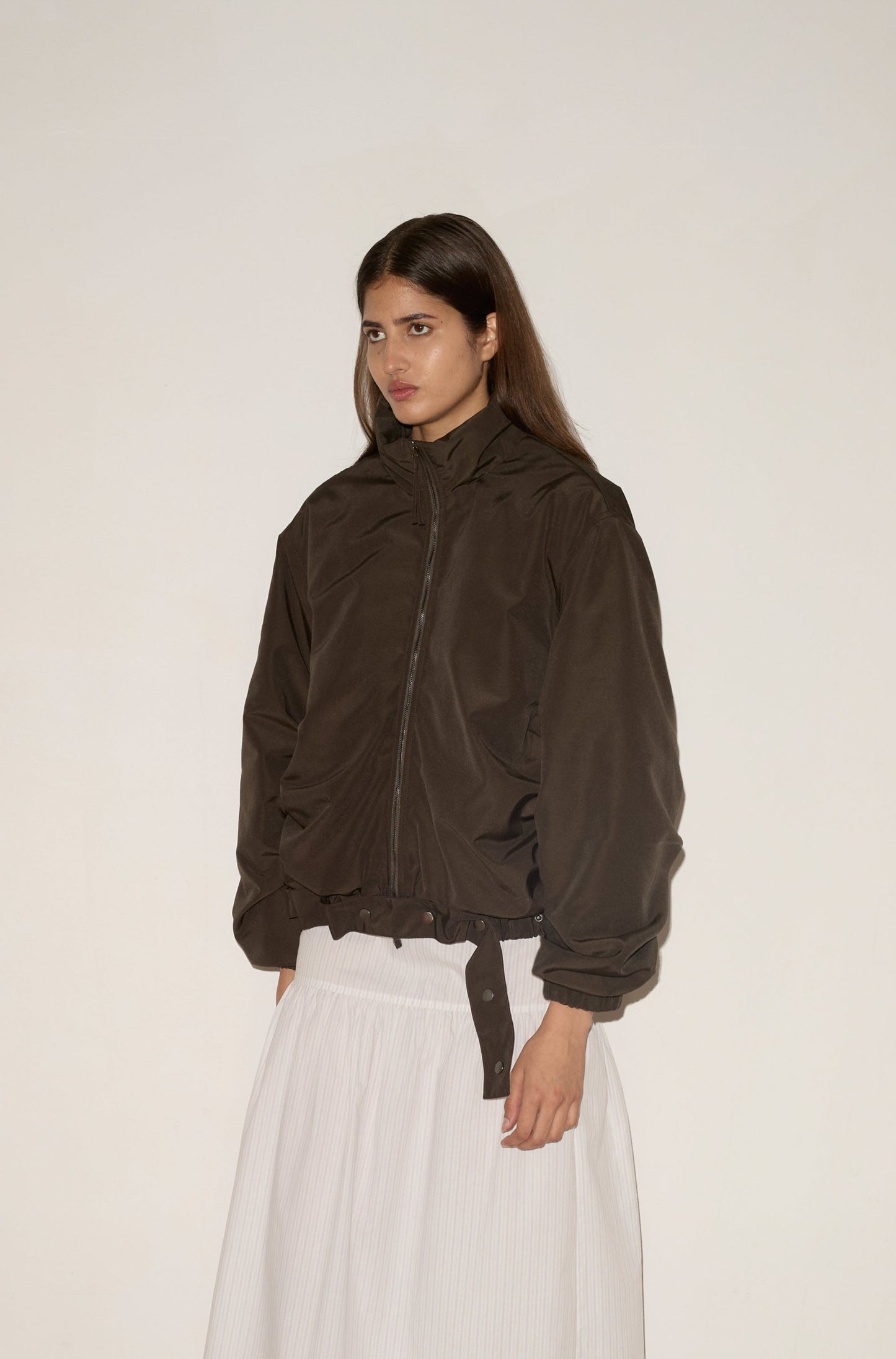 A woman stands facing forward against a plain, light background, wearing the 0 Press Stud Jacket in Dark Chocolate over a long white pleated skirt, her expression neutral.