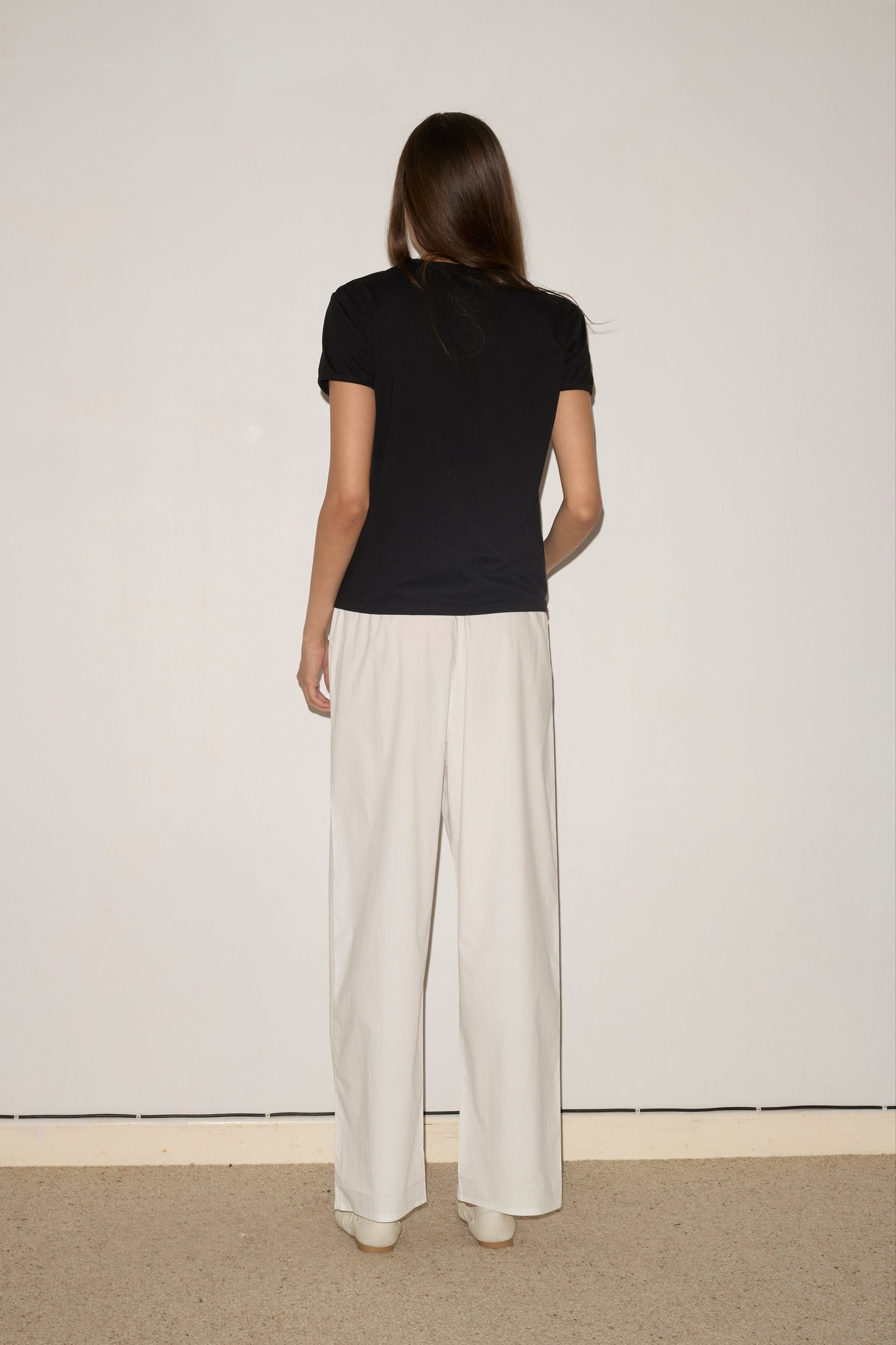 A woman with straight brown hair faces an off-white wall, wearing the 0 Simple Tee - Black and relaxed white pants. The floor beneath her is carpeted in beige.