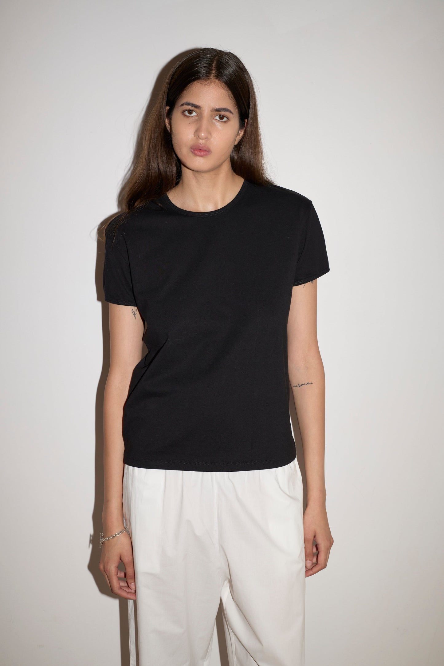 A woman with long brown hair wears the Simple Tee - Black and white pants, standing against a plain white wall while looking at the camera with a neutral expression.