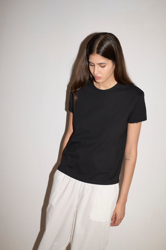 A woman with long brown hair stands against a light background, looking down. She wears the Simple Tee - Black from 0 and white pants, one hand in her pocket and a neutral expression on her face.