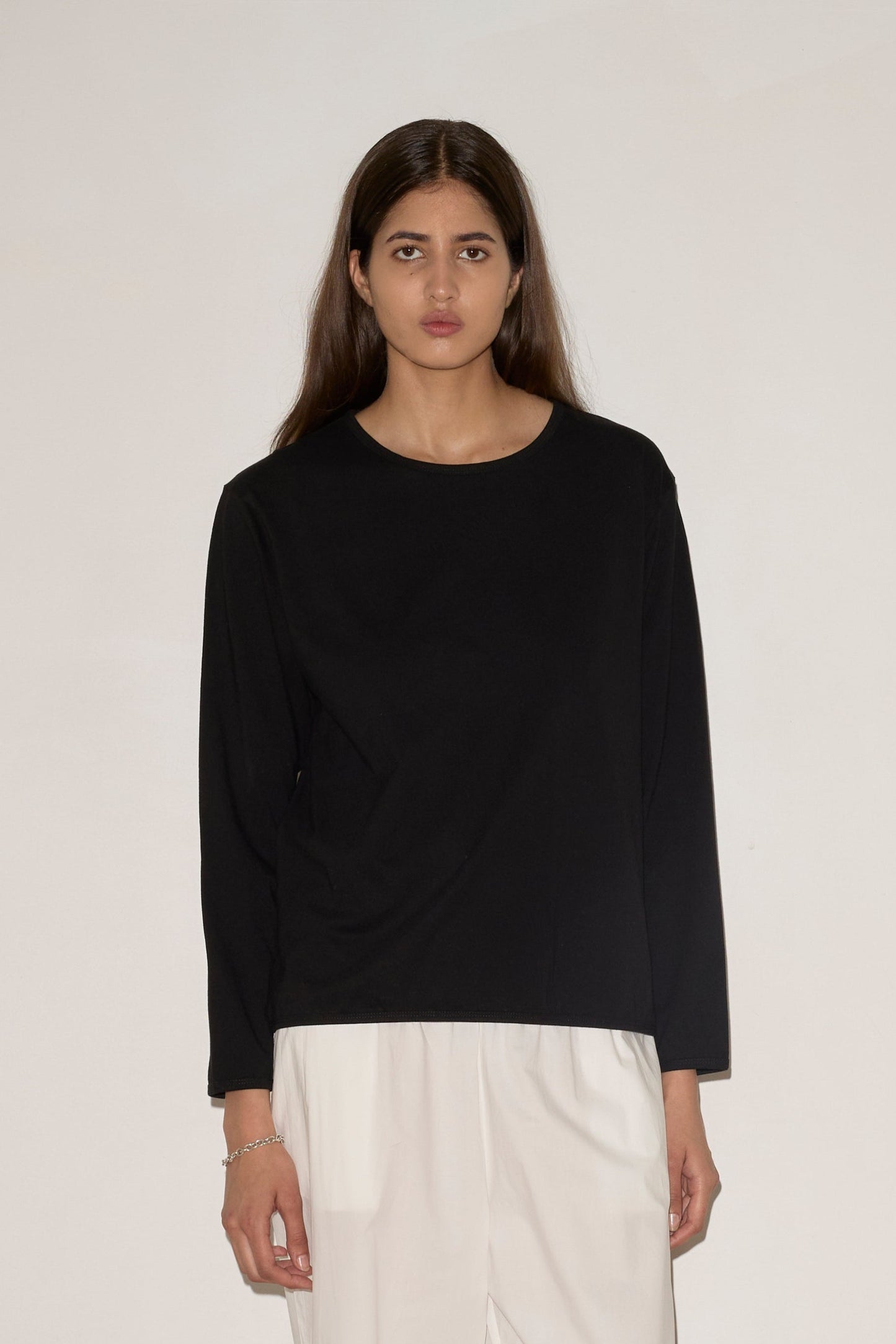 A woman with long brown hair stands against a light wall, wearing a loose black 0 Simple Long Sleeve top, wide white pants, and cream flats. She looks straight ahead with a neutral expression.