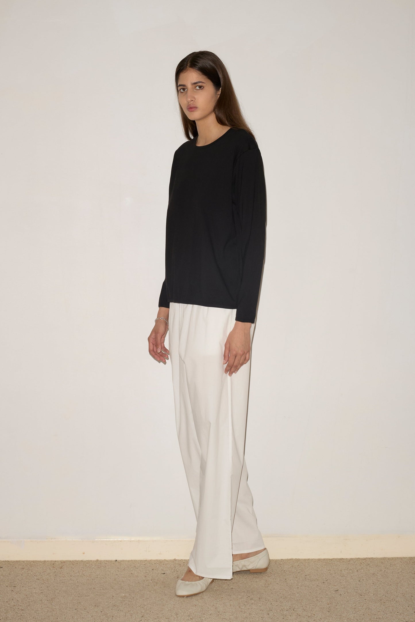 A woman stands indoors against a plain light wall, wearing the 0 Simple Long Sleeve – Black, white wide-leg pants, and beige shoes. She gazes forward with a neutral expression.