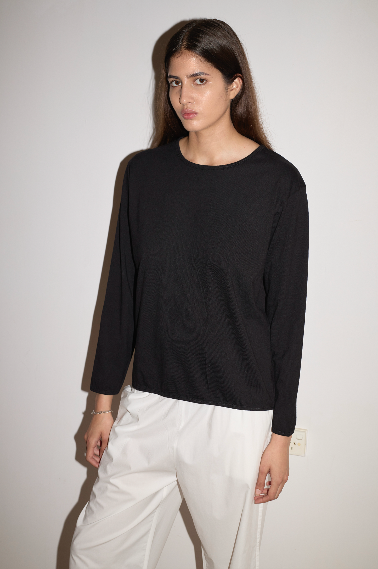 A woman with long brown hair stands against a white wall, wearing the Simple Long Sleeve - Black by 0 and white pants. She faces the camera with a neutral expression.