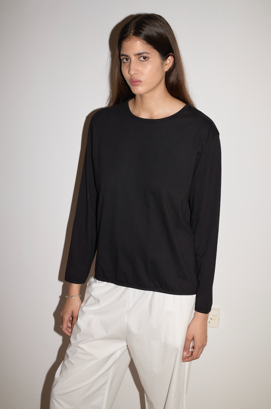 A woman with long brown hair stands against a white wall, wearing the Simple Long Sleeve - Black by 0 and white pants. She faces the camera with a neutral expression.