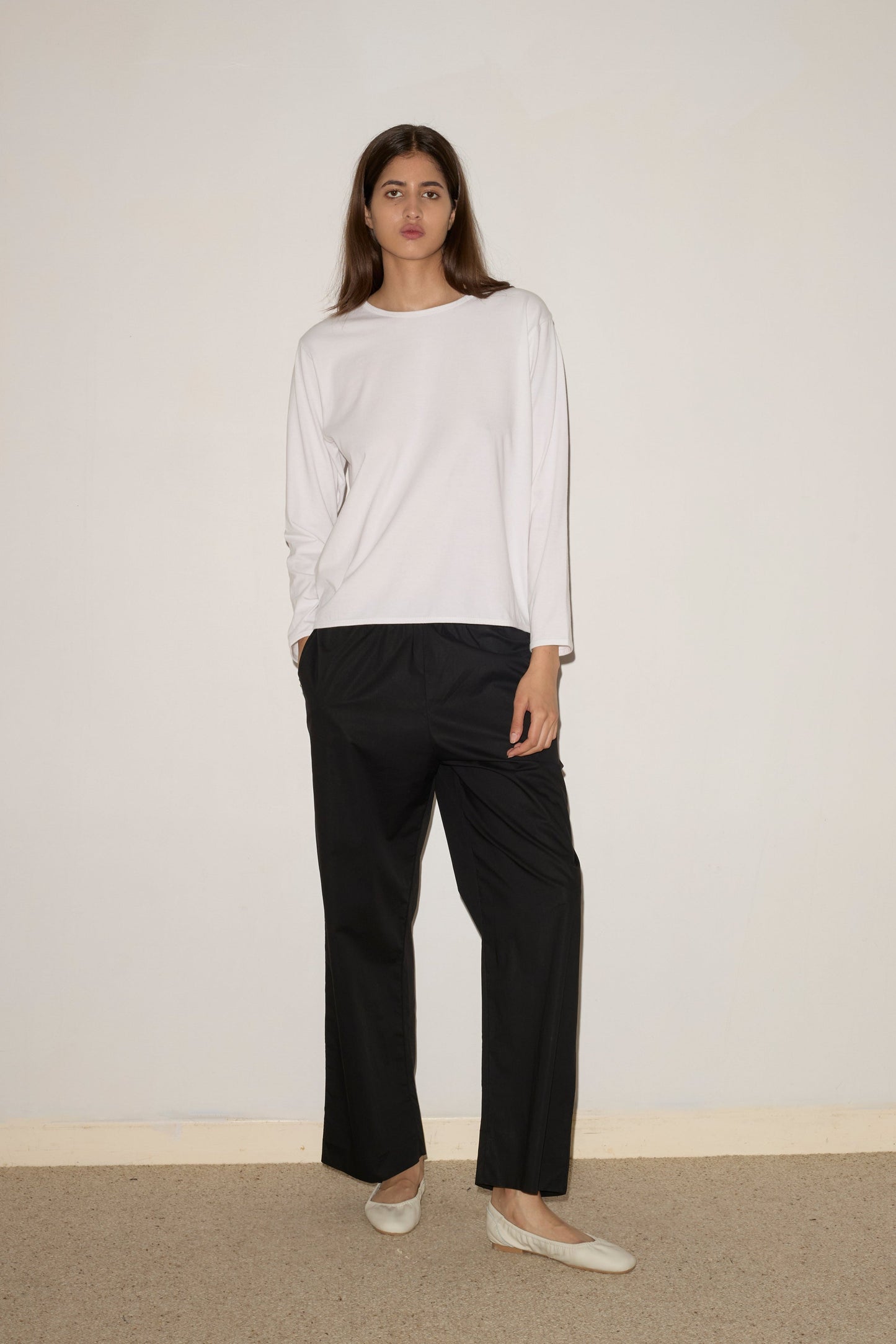 A person with long brown hair stands against a plain light wall, wearing the 0 Simple Long Sleeve in white, black wide-leg pants, and beige slip-on shoes. Their hands are in their pockets and their expression is neutral.