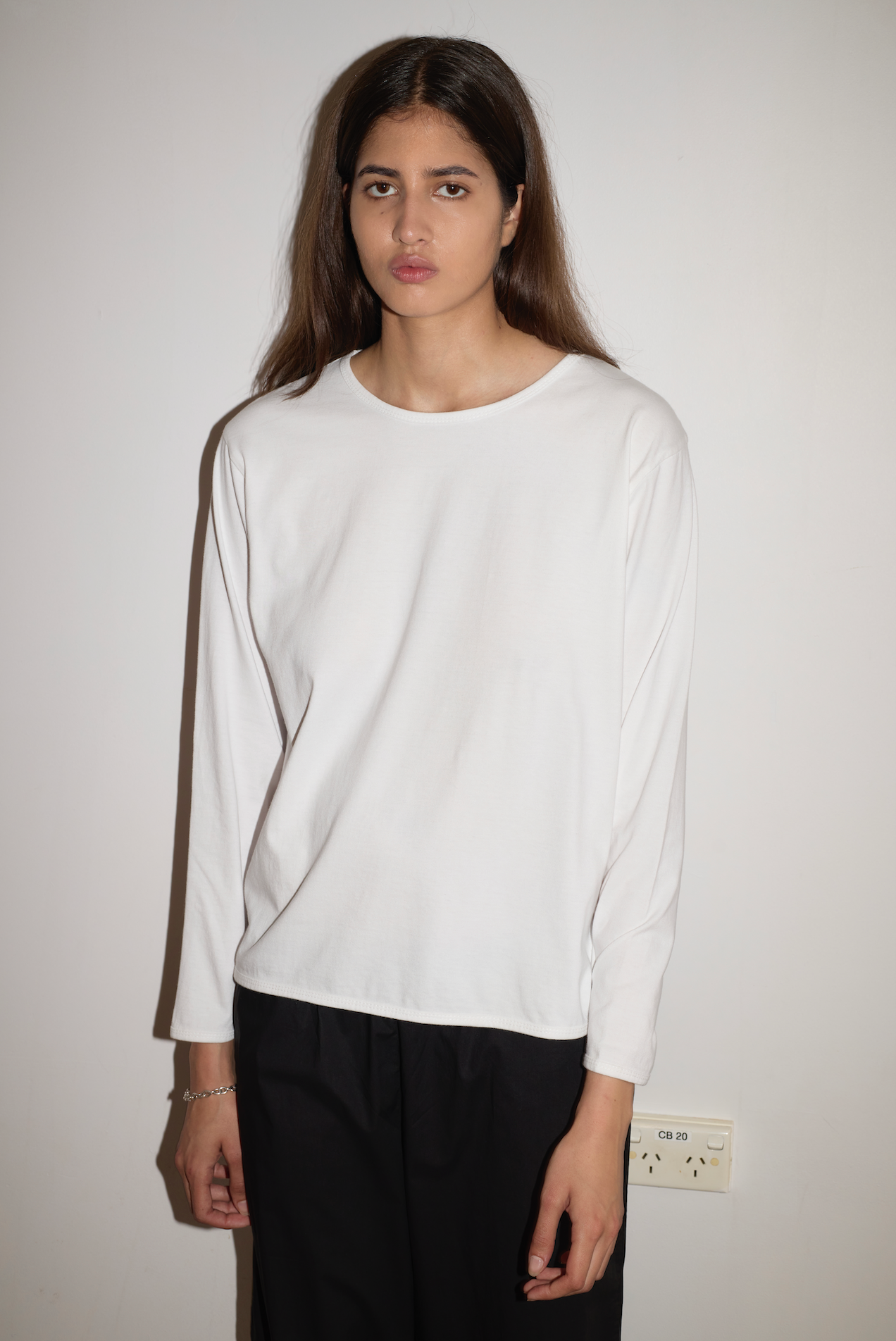 A person with long brown hair wears the 0 Simple Long Sleeve - White shirt and black pants, standing against a white wall with a neutral expression. An electrical outlet is visible on the wall near their hand.