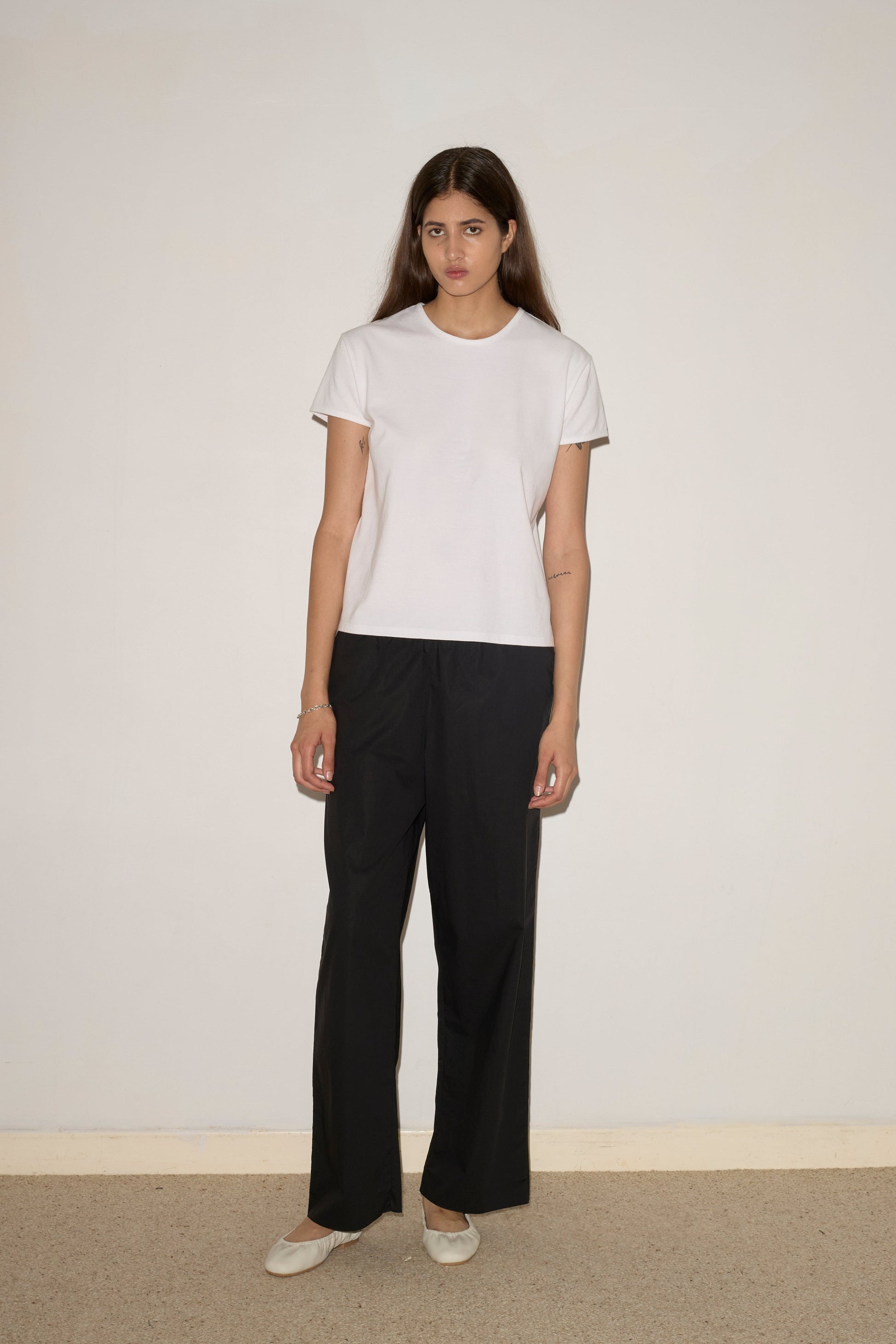 A woman with long brown hair stands against a light wall, wearing the 0 Simple Tee - White (relaxed fit, organic cotton), black pants, and white shoes. Her arms rest at her sides and she has a neutral expression.