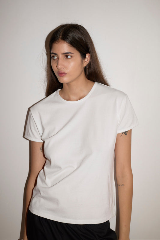 A young woman with long brown hair stands against a plain white wall, wearing the Simple Tee - White by 0 with black pants. She looks slightly to her left and has a neutral expression.