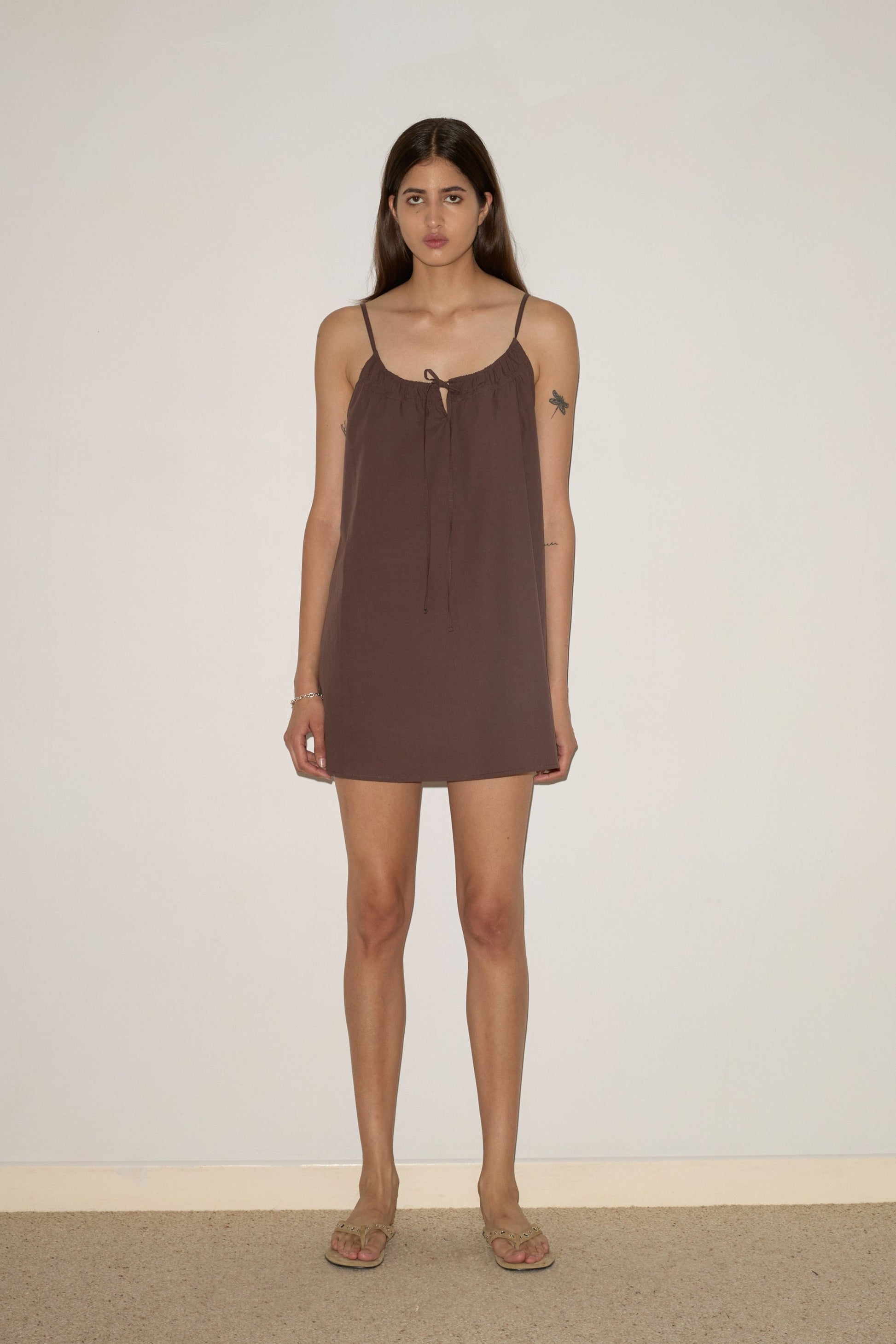 A woman with long brown hair stands by an off-white wall, wearing the Light Dress - Chocolate by 0—a sleeveless mini dress with thin straps—paired with flat sandals. She has a small tattoo on her upper left arm.