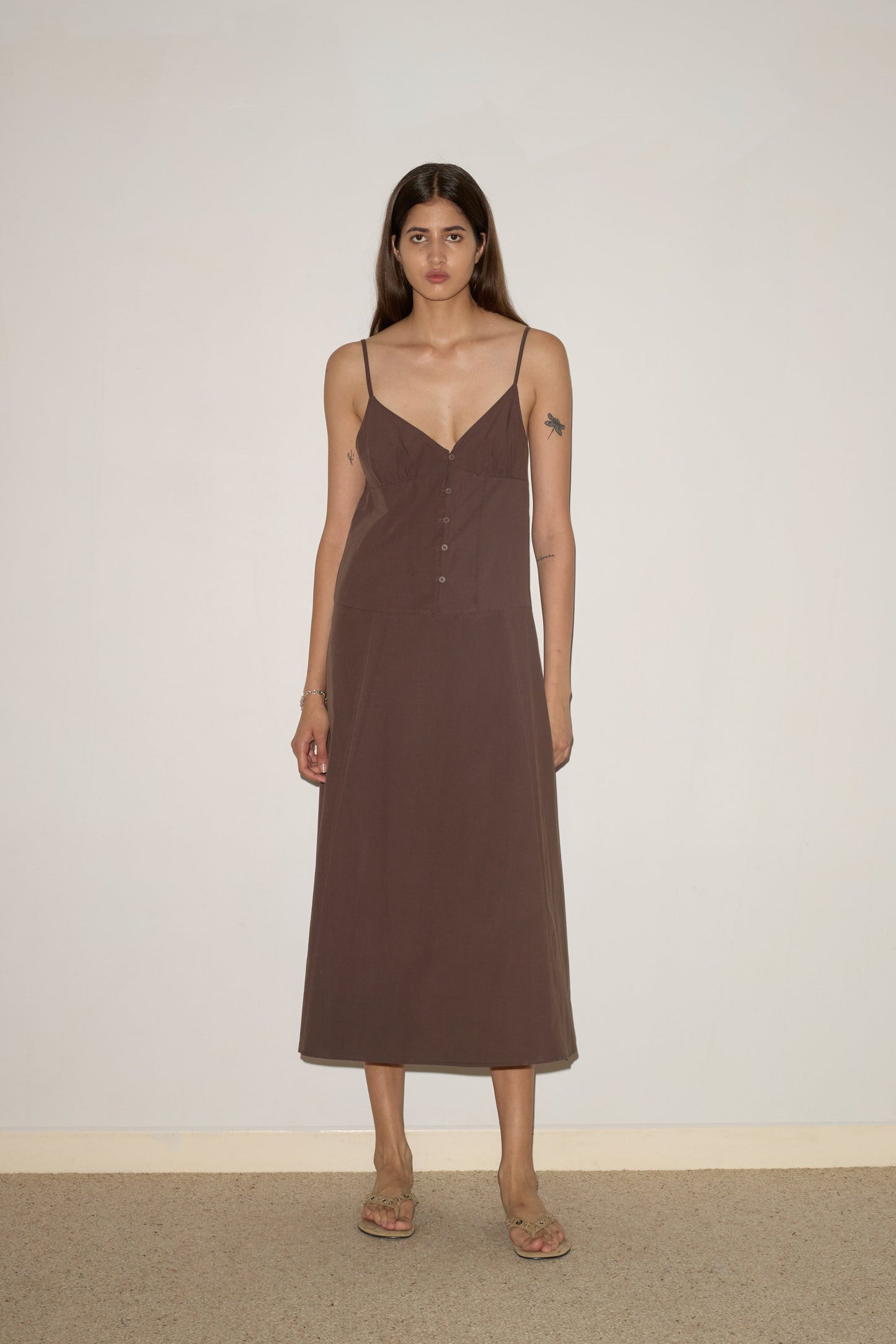 A woman with long brown hair stands indoors against a light wall, wearing the 0 Double Seam Dress - Chocolate, a sleeveless dress with thin straps and front buttons, paired with beige sandals.