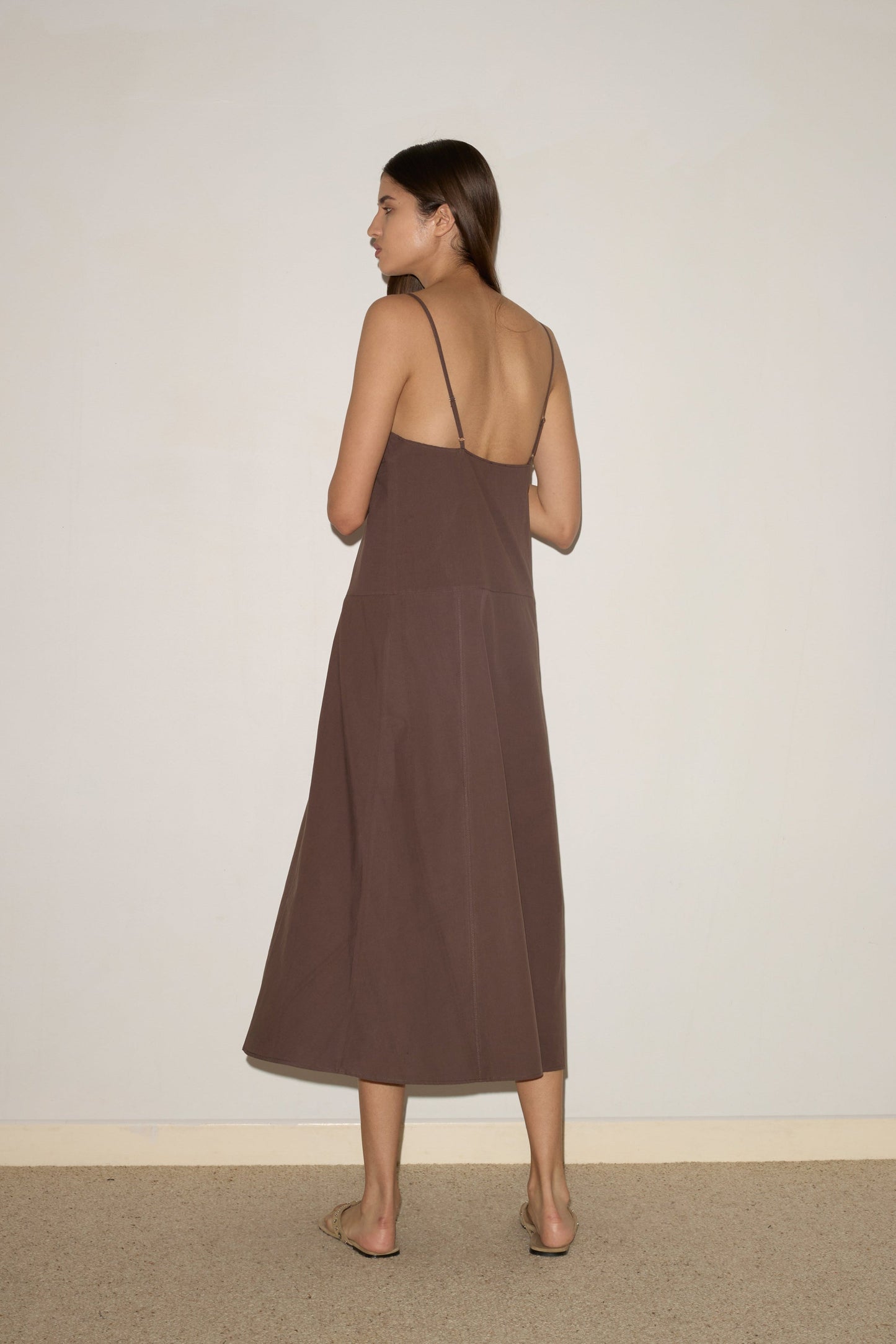 A woman with straight brown hair stands barefoot, facing away from the camera, wearing the 0 Double Seam Dress - Chocolate, an organic cotton sleeveless backless midi with thin straps, against a plain light-colored wall.