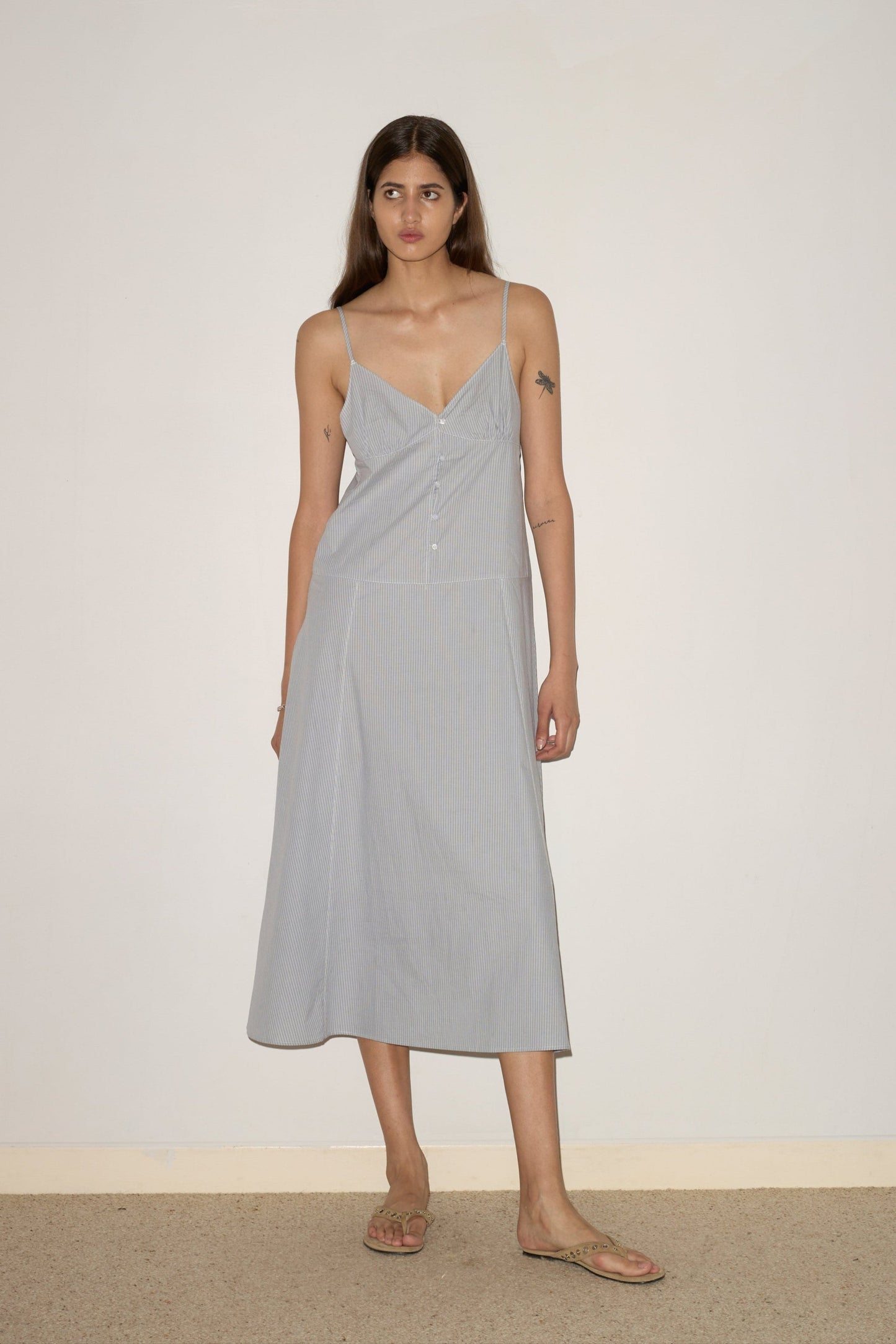 A woman with long brown hair stands against a light wall, barefoot, wearing the Double Seam Dress - Sky Stripe by 0—a light blue midi dress with thin straps and front buttons. She has a small tattoo on her upper arm.