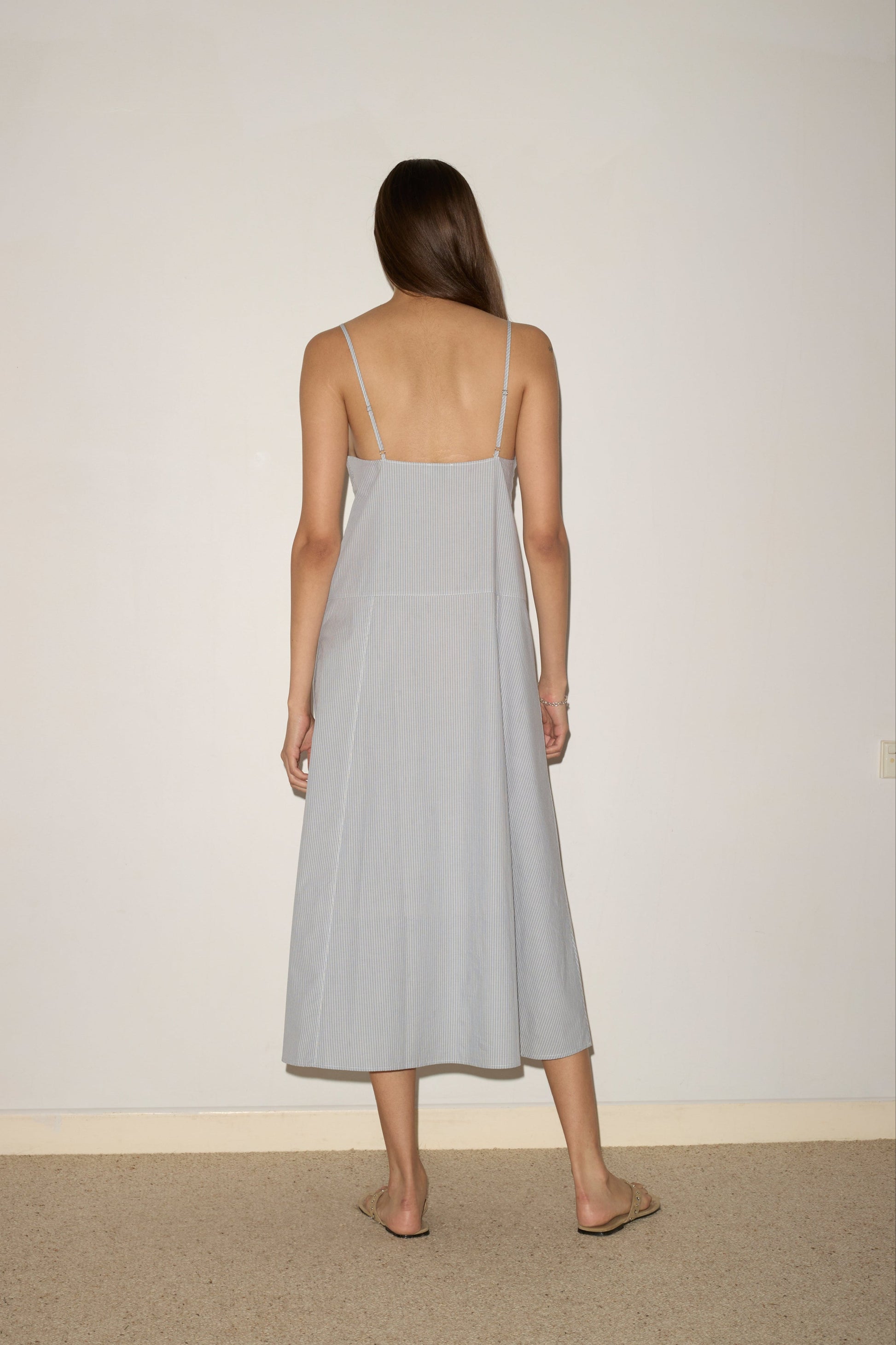 A woman with long brown hair, wearing the Double Seam Dress - Sky Stripe by 0 and sandals, stands facing a plain beige wall with her back to the camera.