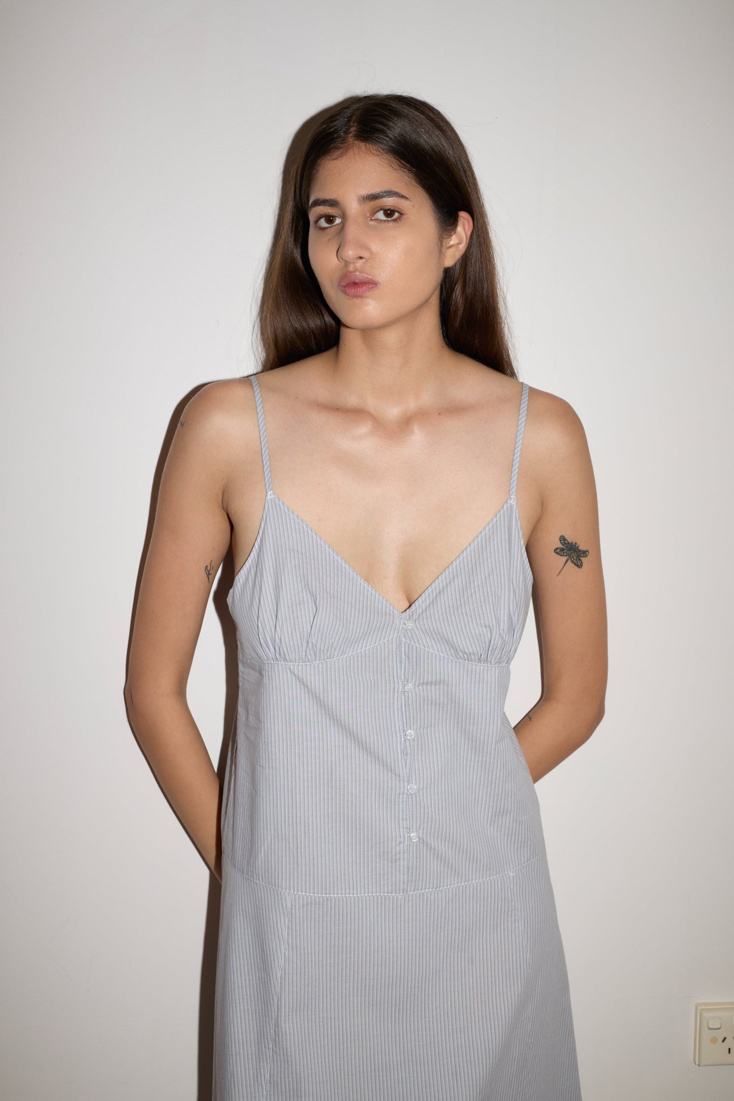 A woman with long brown hair and a dragonfly tattoo on her upper left arm stands against a white wall, wearing the Double Seam Dress - Sky Stripe by 0, featuring thin white stripes. She has a neutral expression.