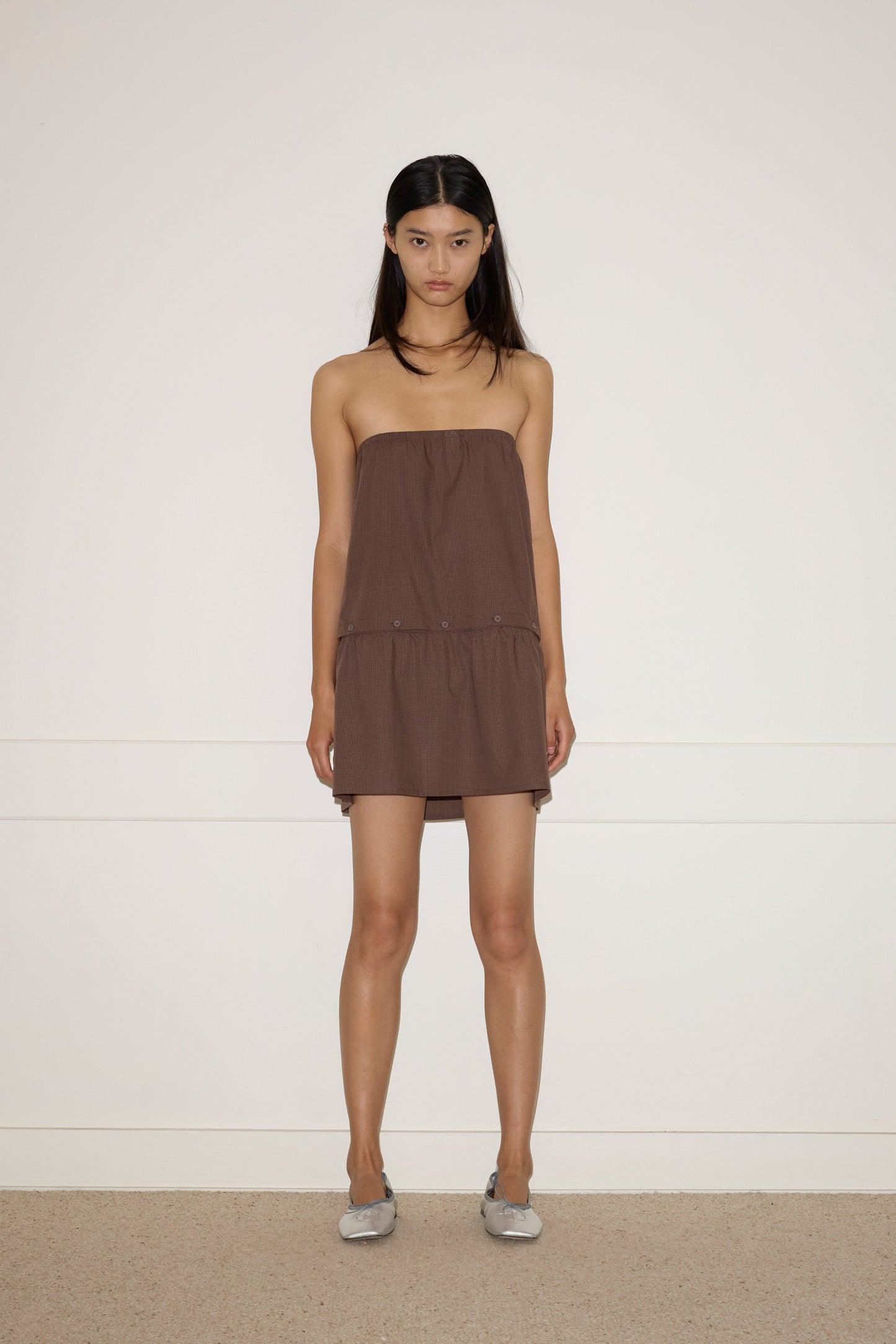 A woman with long dark hair stands against a plain white wall, wearing the Button Dress - Carob Check from Deiji Studios and gray flats. Her arms are relaxed by her sides and she maintains a neutral expression.