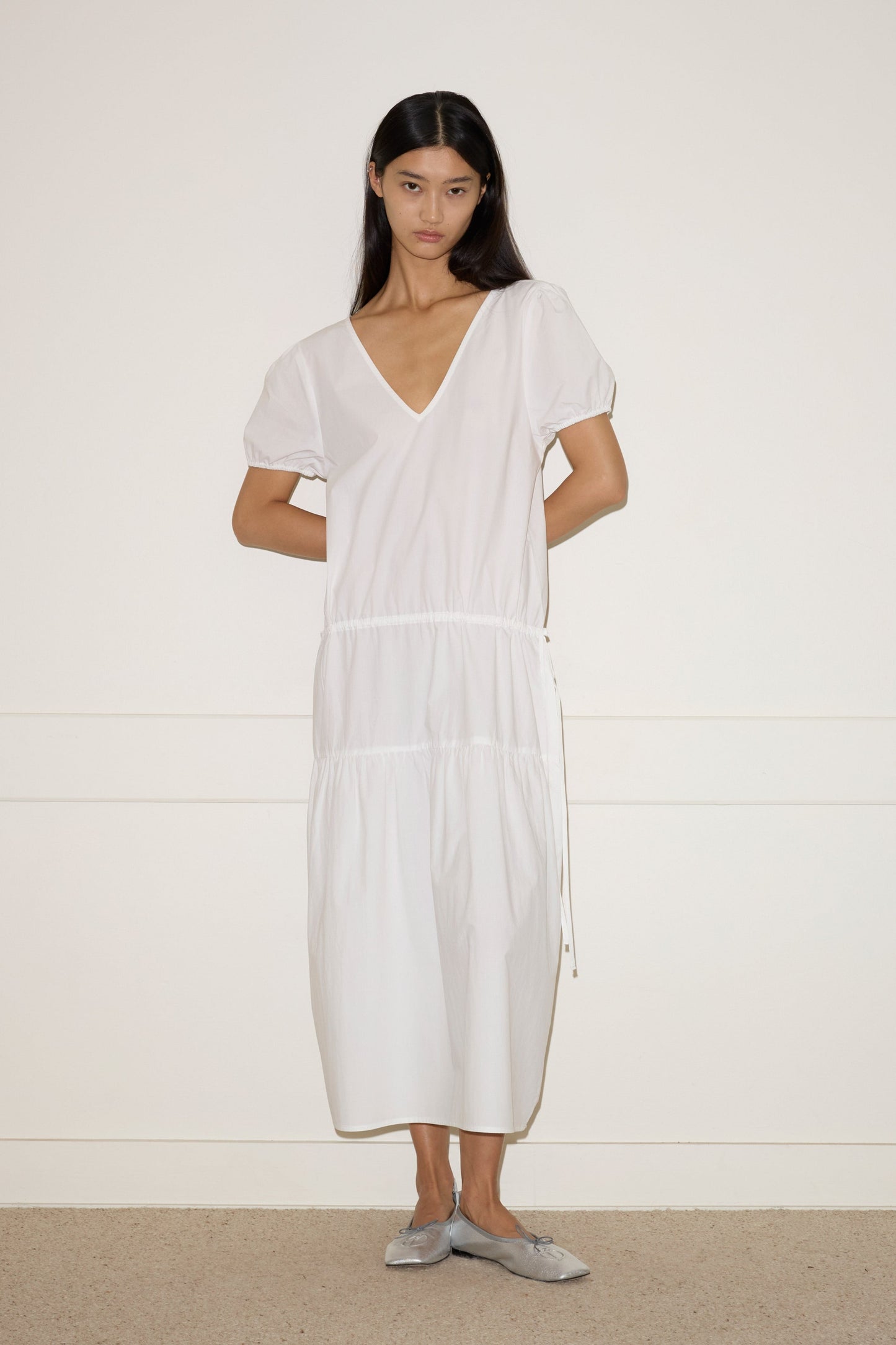 A woman with long dark hair stands against a white wall, wearing the Waistcord Dress—a loose-fitting white V-neck midi dress with short puff sleeves—and gray flat shoes. Her arms are relaxed behind her back.