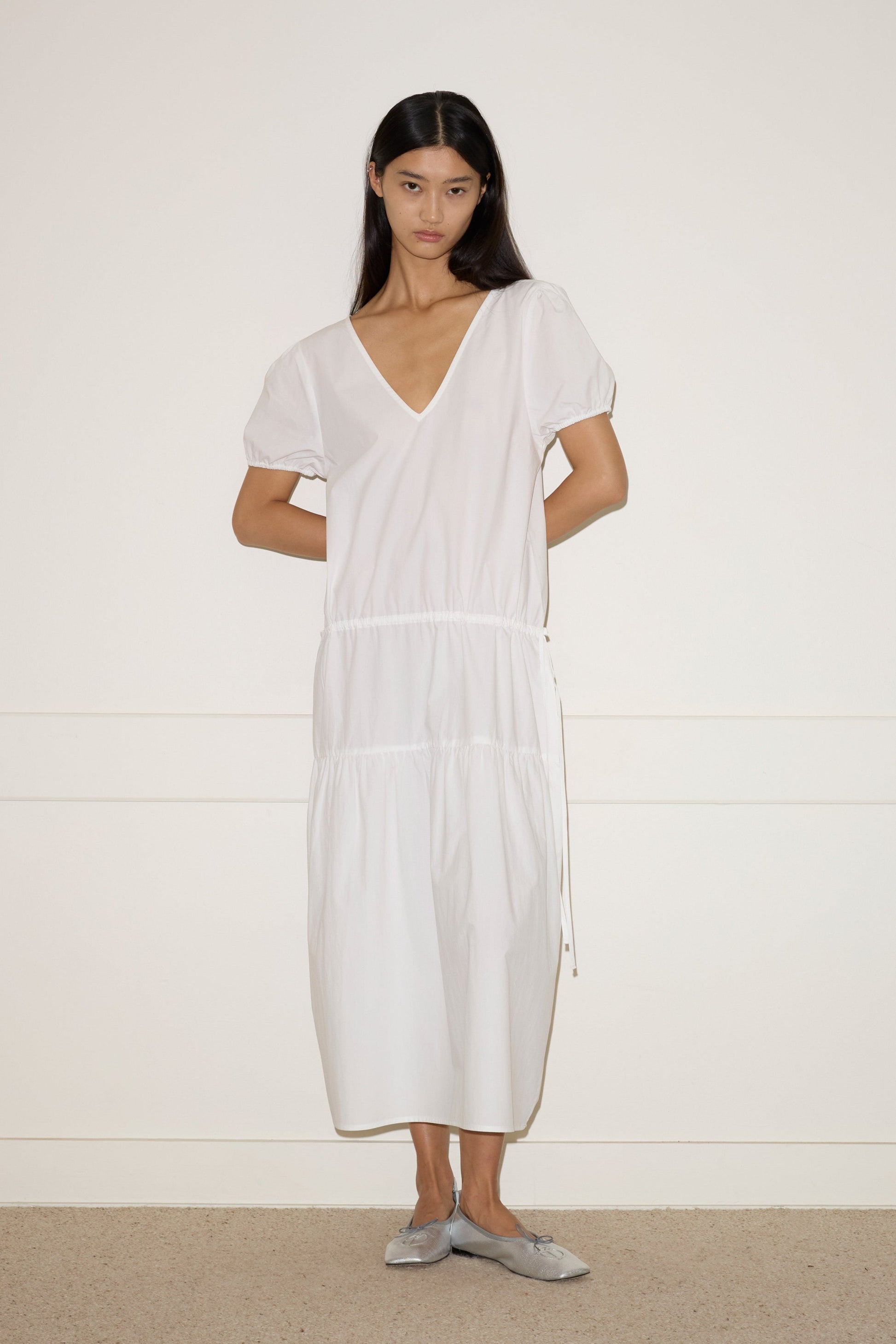 A woman with long dark hair stands against a white wall, wearing the Waistcord Dress—a loose-fitting white V-neck midi dress with short puff sleeves—and gray flat shoes. Her arms are relaxed behind her back.