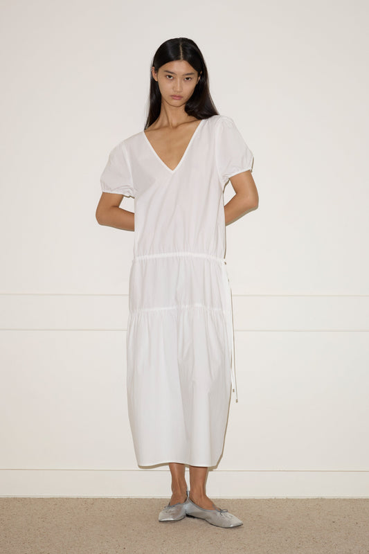 A woman with long dark hair stands against a white wall, wearing the Waistcord Dress—a loose-fitting white V-neck midi dress with short puff sleeves—and gray flat shoes. Her arms are relaxed behind her back.