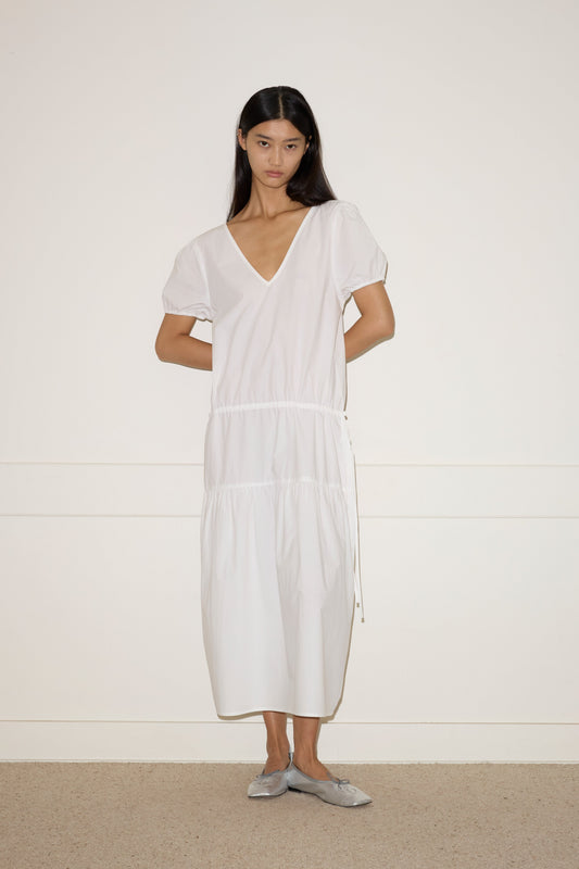 A woman with long dark hair stands against a white wall, wearing the Waistcord Dress—a loose-fitting white V-neck midi dress with short puff sleeves—and gray flat shoes. Her arms are relaxed behind her back.