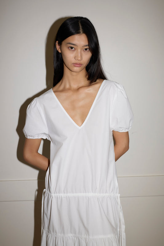 A person with long black hair stands against a light wall, hands behind their back, wearing the white Waistcord Dress by Deiji Studios—a loose, short-sleeved organic cotton dress with a deep V-neckline.