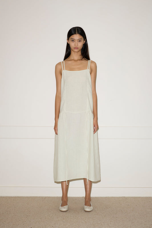 A woman with long dark hair stands against a plain wall, wearing the Linen Strap Dress—a light beige, sleeveless, loose-fitting midi dress with thin straps—paired with flats. She faces forward with a neutral expression.