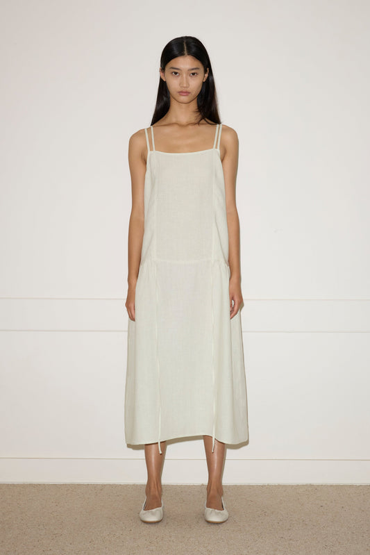 A woman with long dark hair stands against a plain wall, wearing the Linen Strap Dress—a light beige, sleeveless, loose-fitting midi dress with thin straps—paired with flats. She faces forward with a neutral expression.