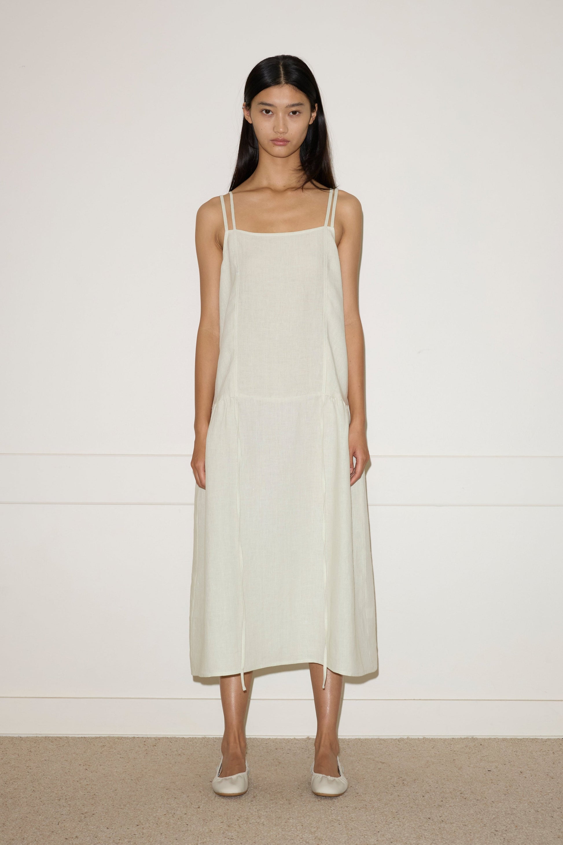 A woman with long dark hair stands against a plain wall, wearing the Linen Strap Dress—a light beige, sleeveless, loose-fitting midi dress with thin straps—paired with flats. She faces forward with a neutral expression.