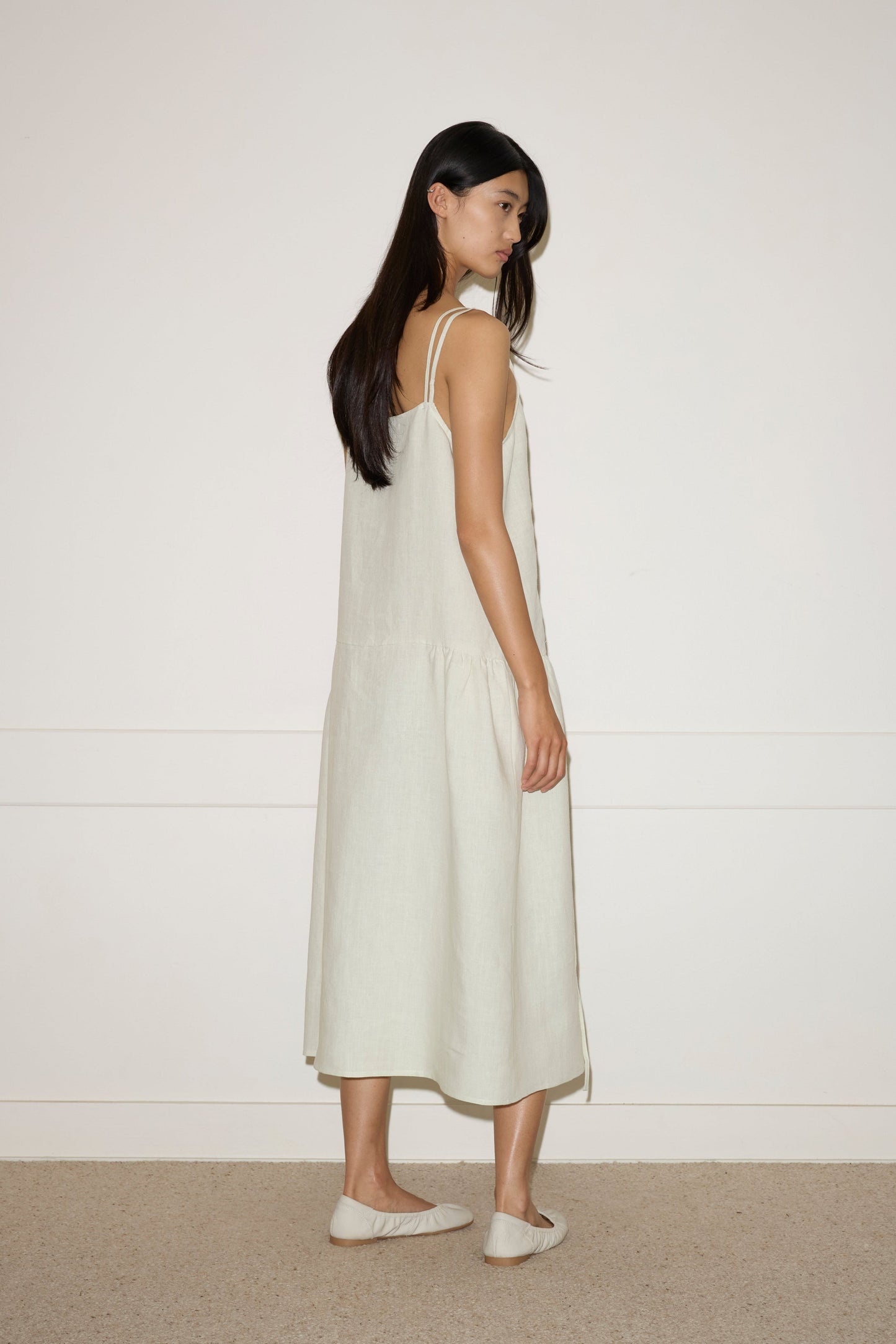A woman with long dark hair stands sideways, wearing a Linen Strap Dress in Pastel Pistachio by Deiji Studios with matching flats, set against a plain white wall and carpeted floor.