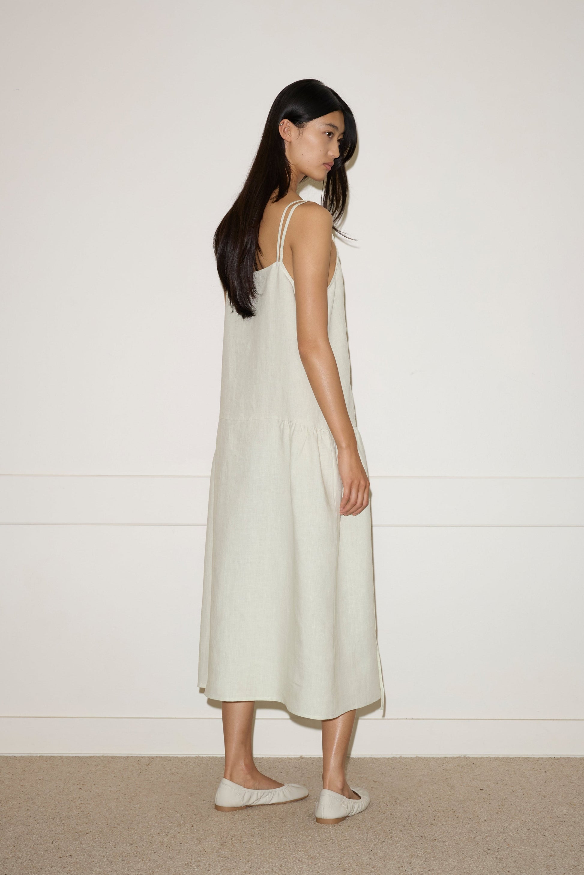 A woman with long dark hair stands sideways, wearing a Linen Strap Dress in Pastel Pistachio by Deiji Studios with matching flats, set against a plain white wall and carpeted floor.