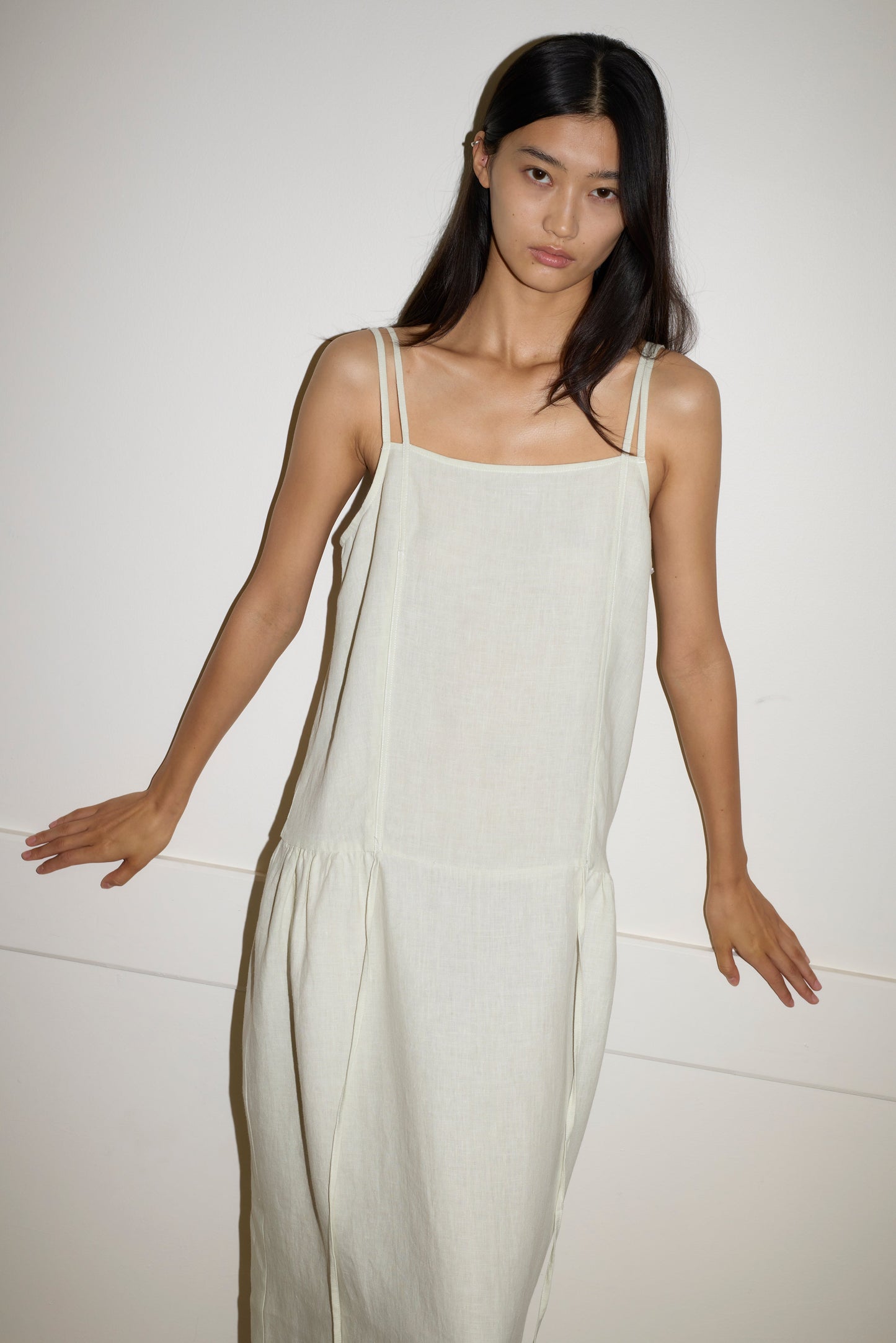 A woman with long dark hair wears a loose-fitting, sleeveless Linen Strap Dress in Pastel Pistachio by Deiji Studios, standing against a plain white wall with her arms slightly extended to the sides.