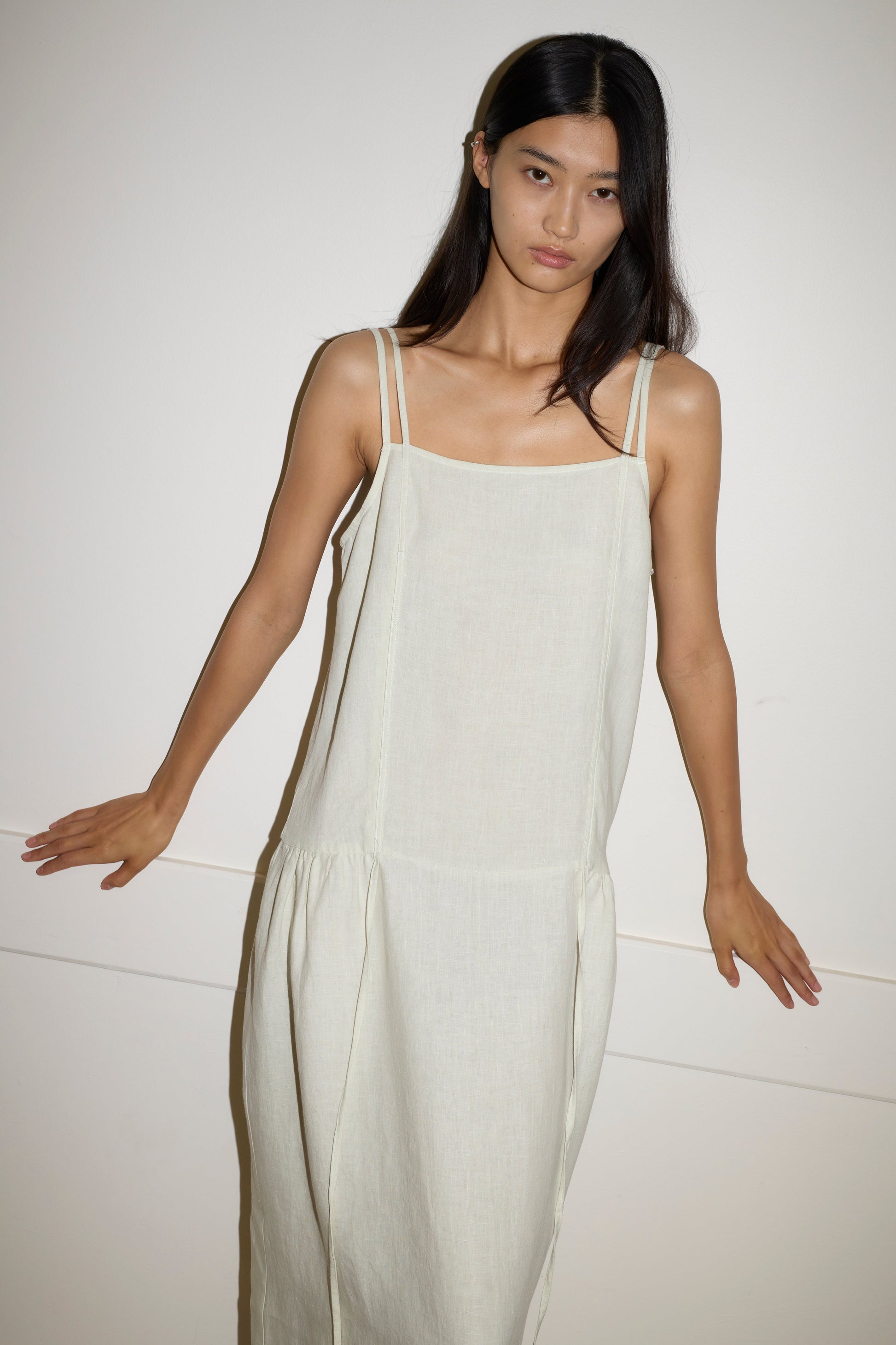 A woman with long dark hair wears a loose-fitting, sleeveless Linen Strap Dress in Pastel Pistachio by Deiji Studios, standing against a plain white wall with her arms slightly extended to the sides.