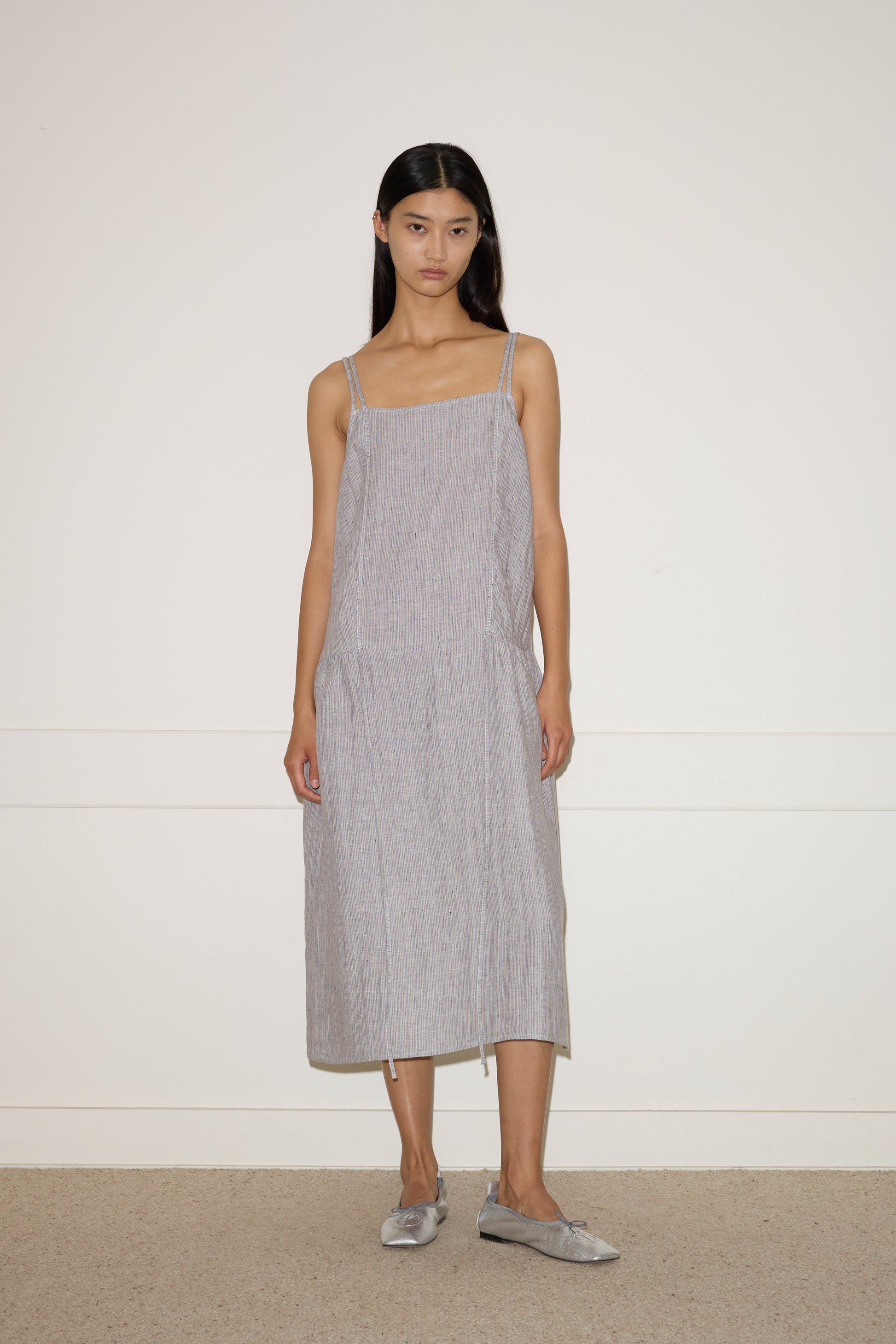 A woman with long dark hair stands against a plain wall, facing forward in a neutral expression. She wears the 0 Linen Strap Dress - Haze Blue, made from stone-washed French linen, paired with matching slip-on shoes.