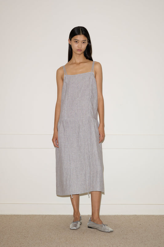 A woman with long dark hair stands against a plain wall, facing forward in a neutral expression. She wears the 0 Linen Strap Dress - Haze Blue, made from stone-washed French linen, paired with matching slip-on shoes.