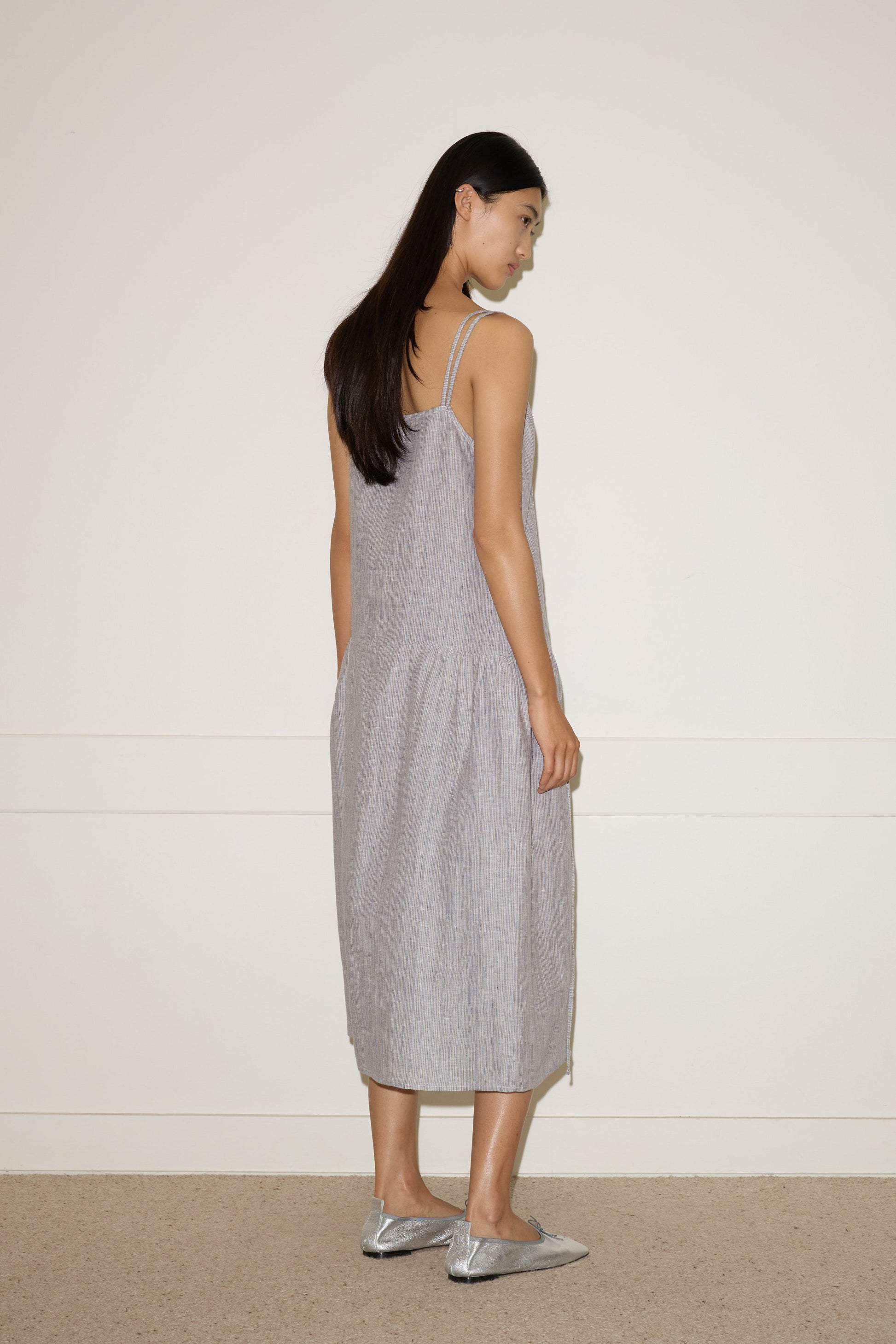 A woman with long dark hair stands sideways, looking back, wearing the 0 Linen Strap Dress - Haze Blue and shiny silver flats, set against a plain light-colored wall and carpeted floor.