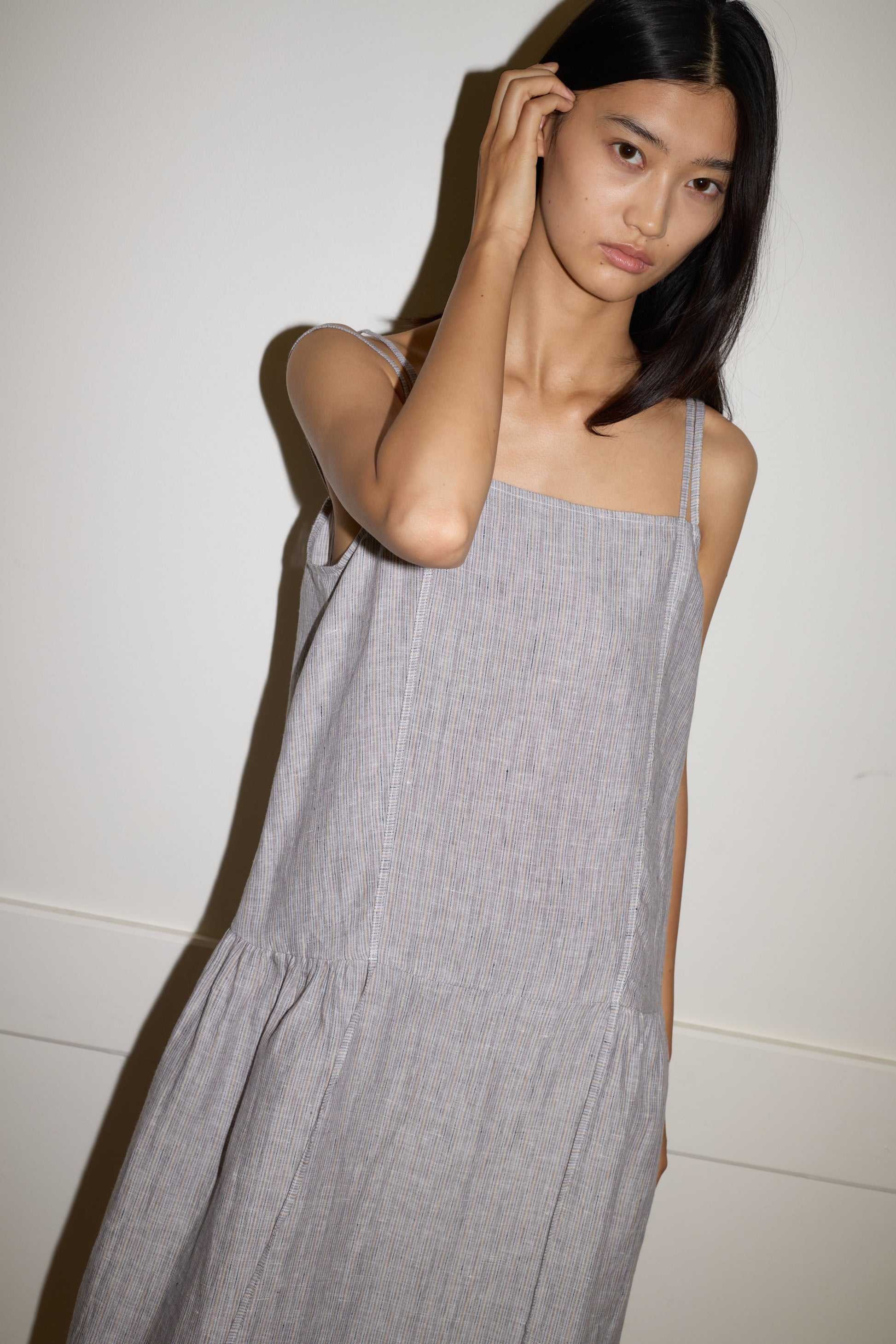 A woman with long dark hair models the Linen Strap Dress - Haze Blue by 0, made from stone-washed French linen. She stands against a white wall, hand at her head, gazing thoughtfully at the camera.