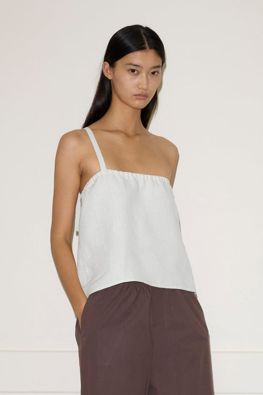 A woman stands indoors by a light wall, wearing a white Button Strap Top - White from Deiji Studios, brown knee-length shorts, and beige flats. She has long straight hair and keeps her hands in her pockets.