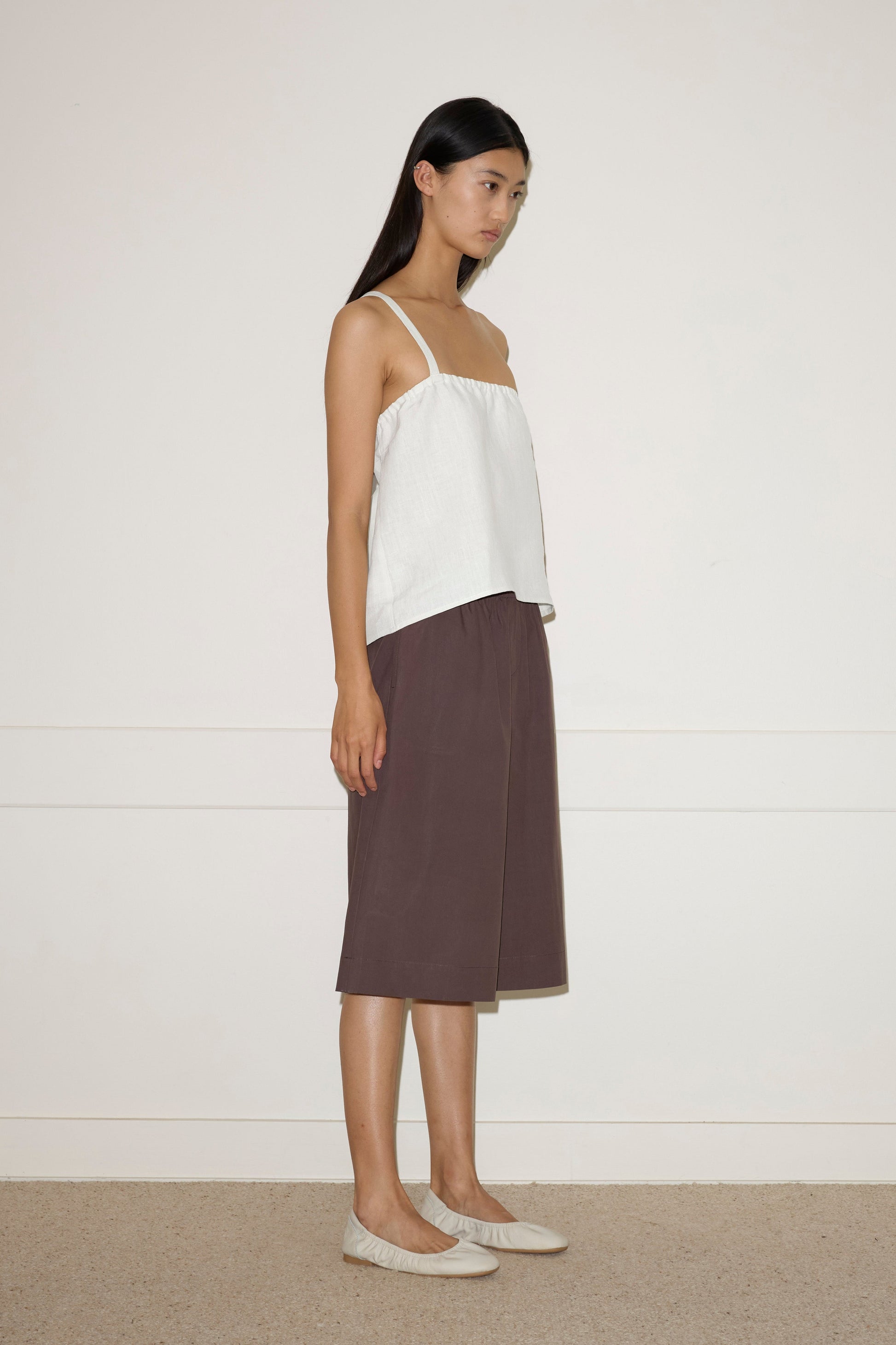 A woman with long dark hair stands sideways against a plain light wall, wearing a loose Button Strap Top - White, brown knee-length shorts, and beige flats.