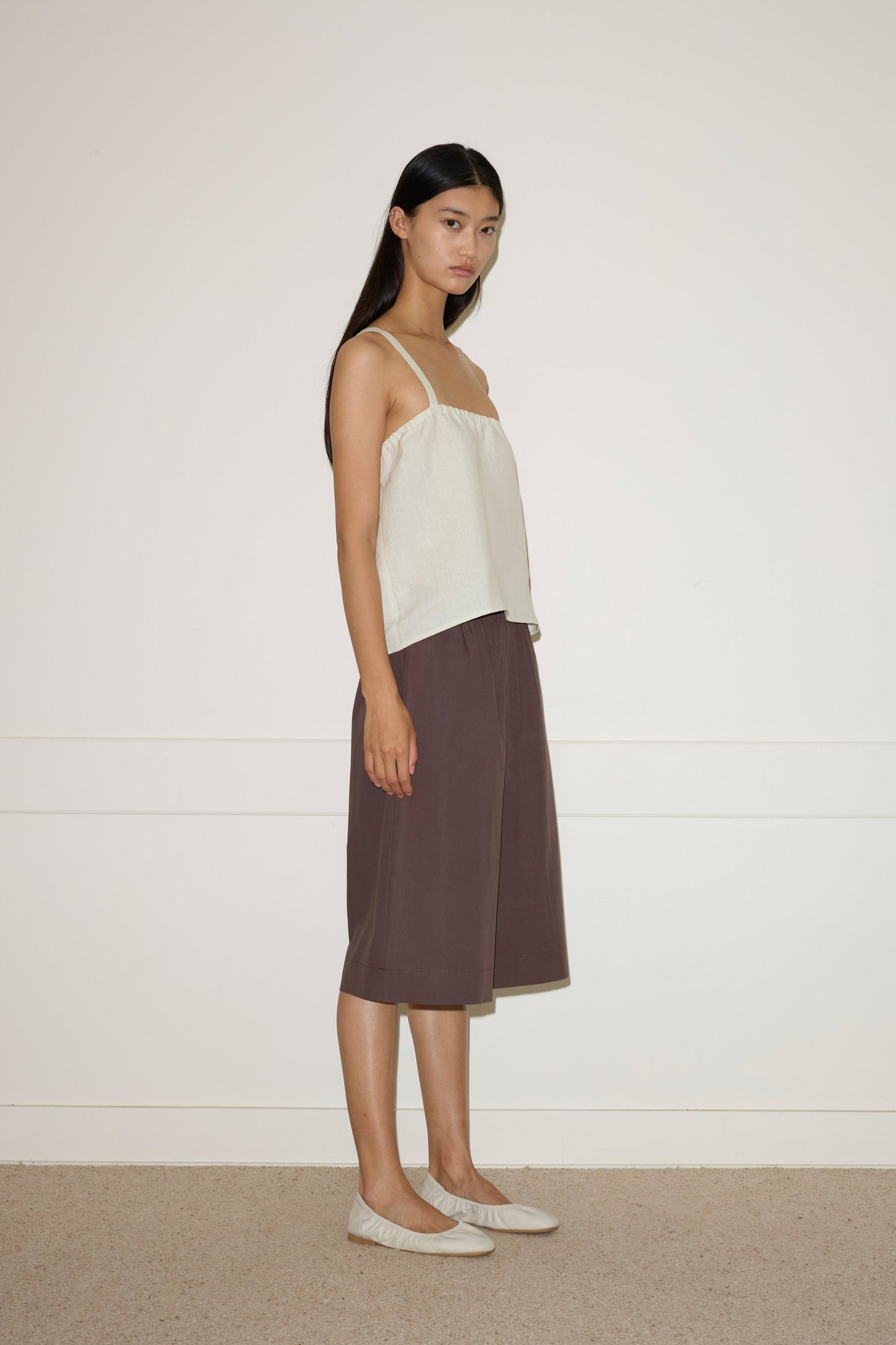 A woman with long dark hair stands sideways against a white wall, wearing the 0 Long Lounger - Chocolate, a relaxed-fit, knee-length brown skirt paired with a light sleeveless top and light flat shoes.