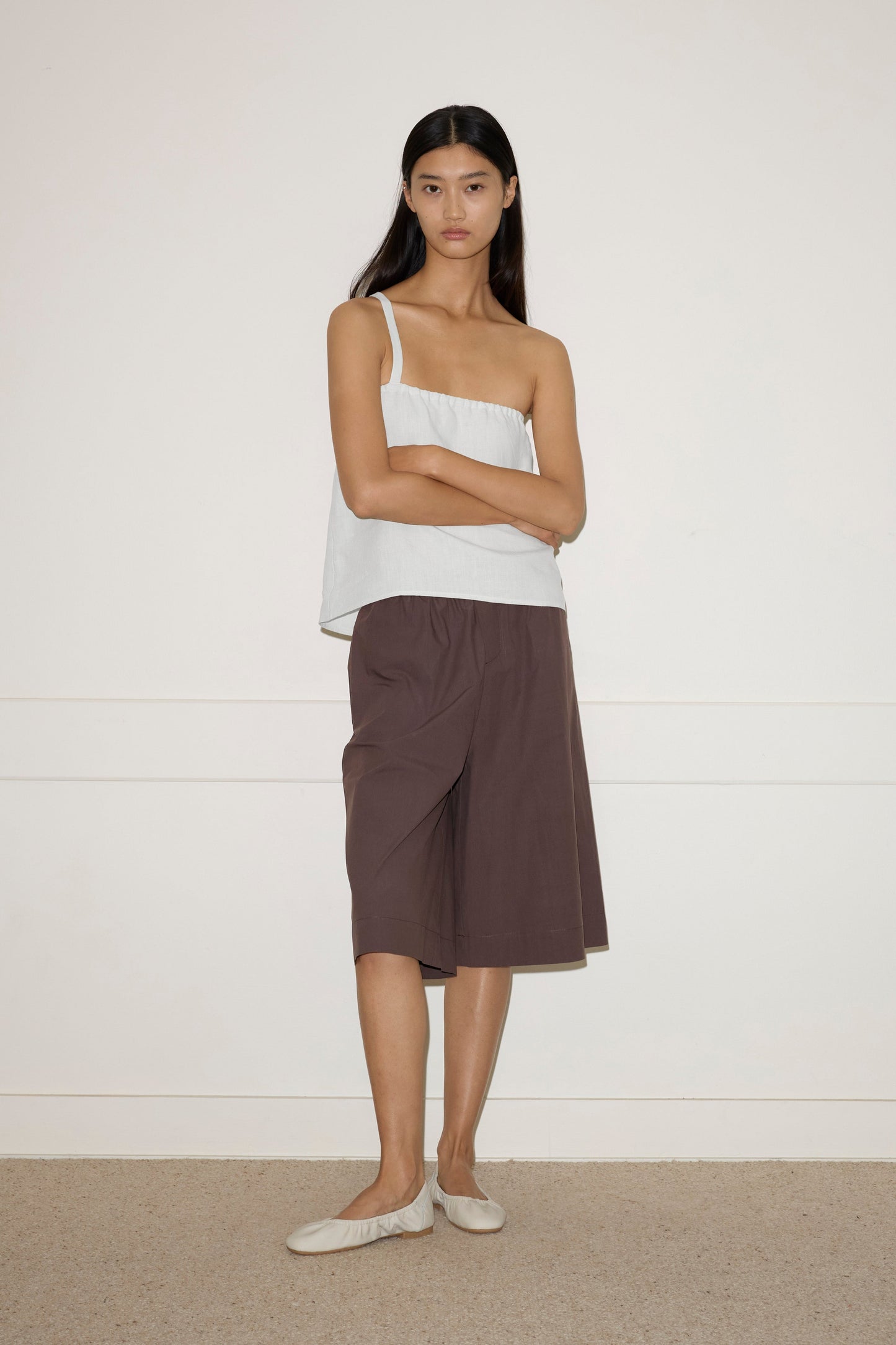A woman with long dark hair stands against an off-white wall, wearing the Button Strap Top - White by Deiji Studios, loose brown knee-length shorts, and beige flats. She crosses her arms and looks straight ahead.