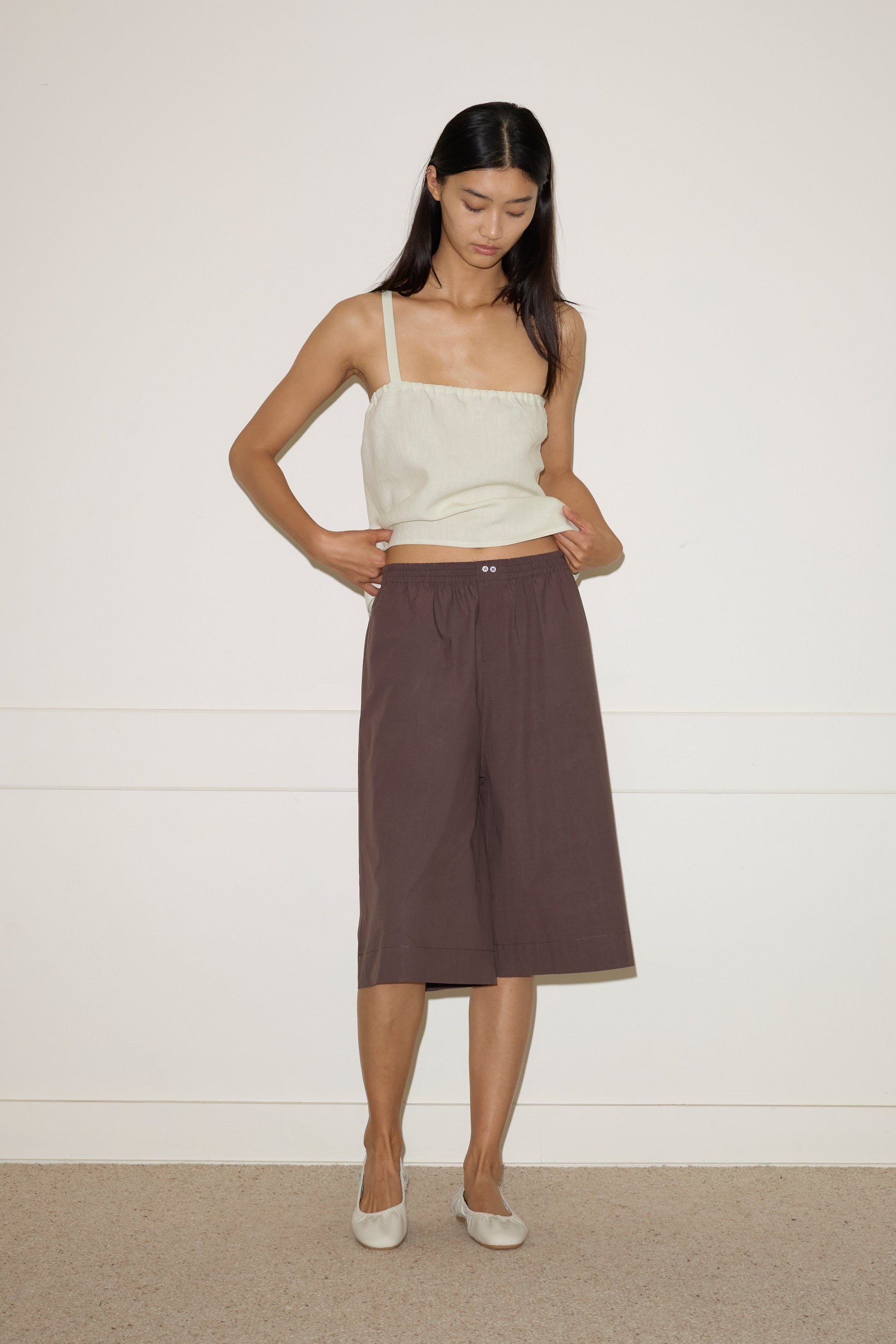 A woman stands against a white wall wearing the Long Lounger - Chocolate by 0, a relaxed-fit, dark knee-length sleeveless piece. She looks down with her hands in her pockets and pairs it with light-colored flat shoes.