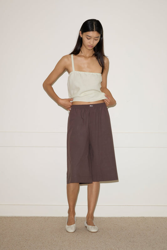 A woman stands against a white wall wearing the Long Lounger - Chocolate by 0, a relaxed-fit, dark knee-length sleeveless piece. She looks down with her hands in her pockets and pairs it with light-colored flat shoes.