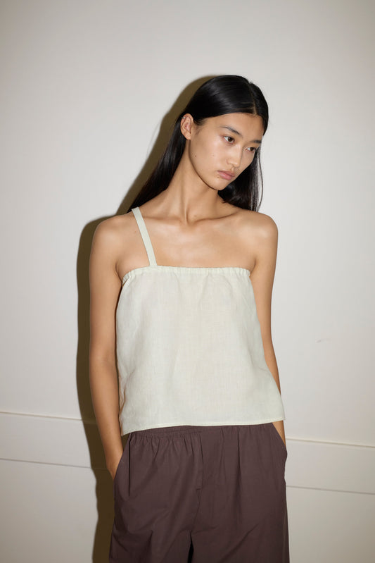 A woman with long, straight black hair wears the Button Strap Top in light beige and loose brown pants, standing against an off-white wall with her hands in her pockets, looking slightly downward.