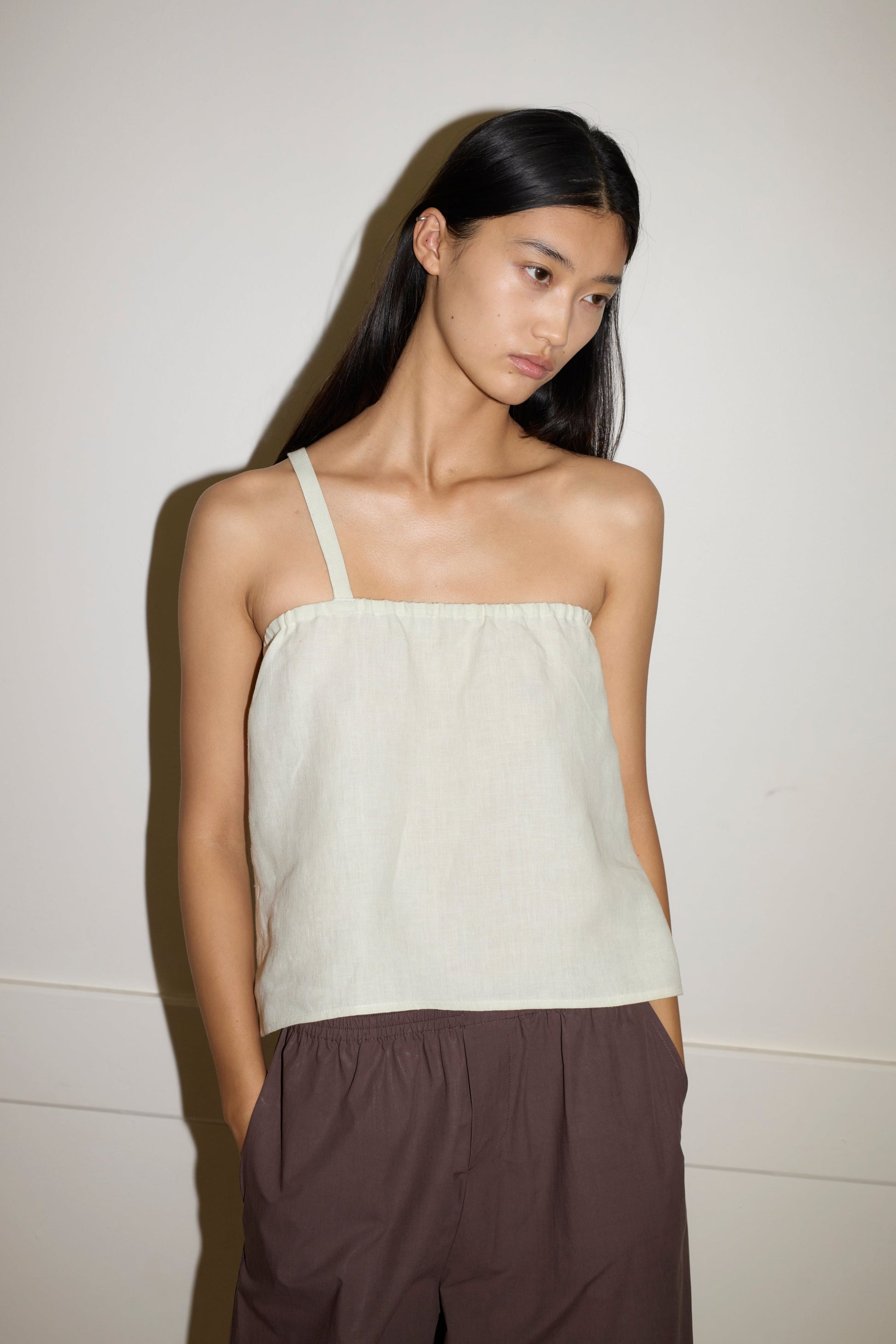 A woman with long, straight black hair wears the Button Strap Top in light beige and loose brown pants, standing against an off-white wall with her hands in her pockets, looking slightly downward.