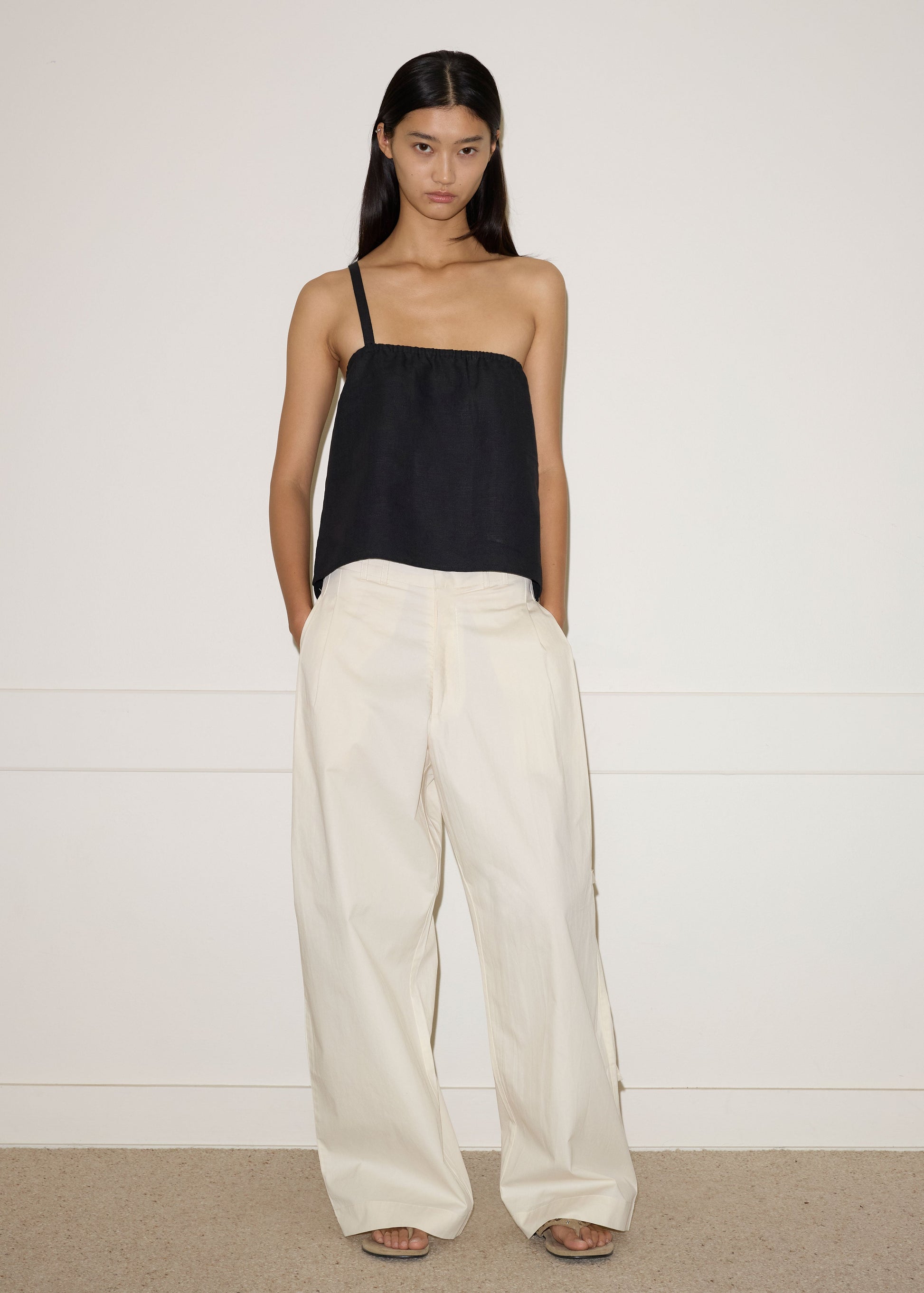 A woman stands indoors against a plain white wall, wearing the Tab Pant in Off White with a loose black sleeveless top. Her hands are in her pockets, she has straight dark hair, and she pairs the look with sandals for a relaxed vibe.