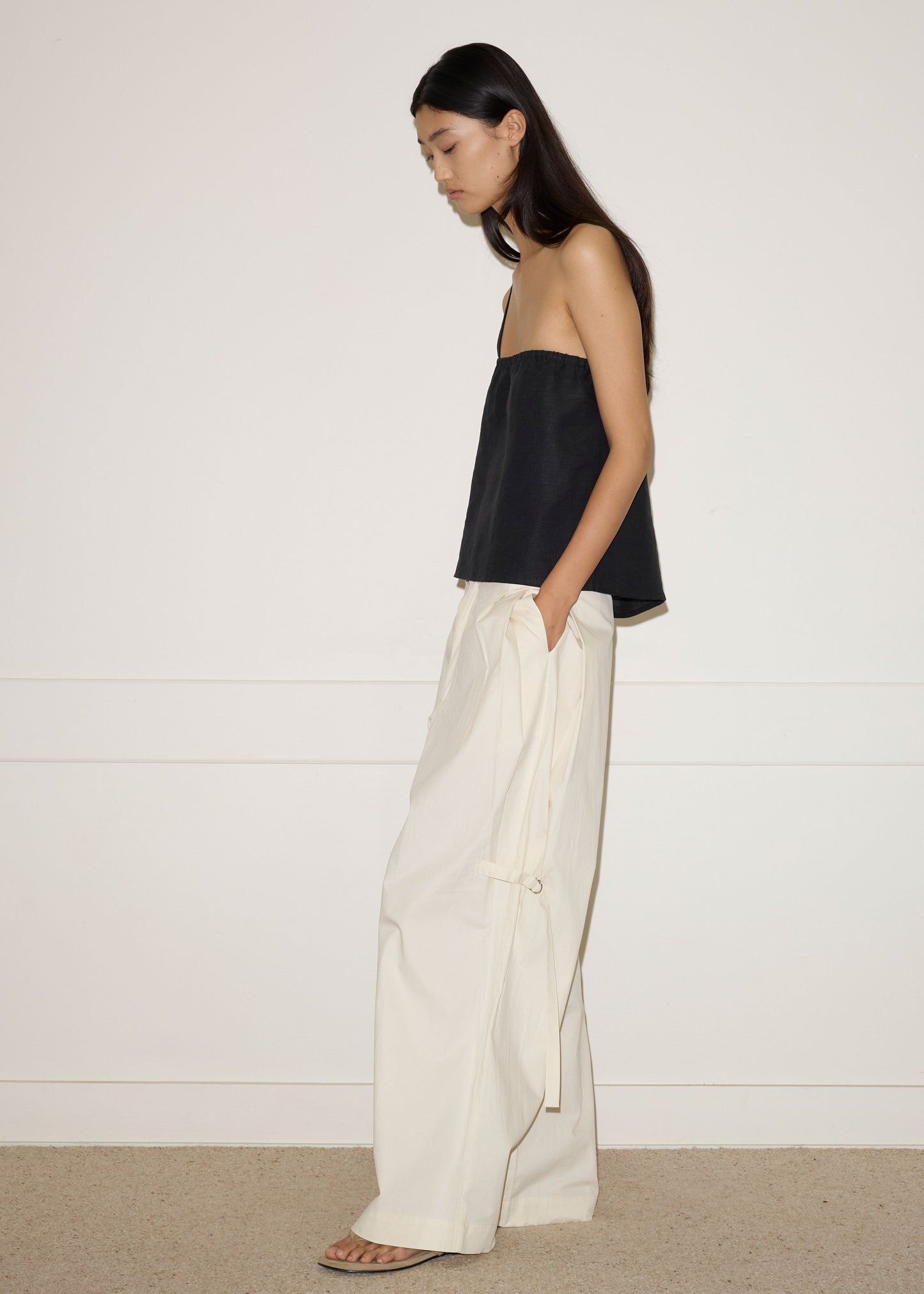 A woman with long dark hair stands in profile against a plain wall, wearing a strapless black top, Deiji Studios Tab Pant in Off White organic cotton, and sandals. One hand rests in her pocket as she looks downwards.