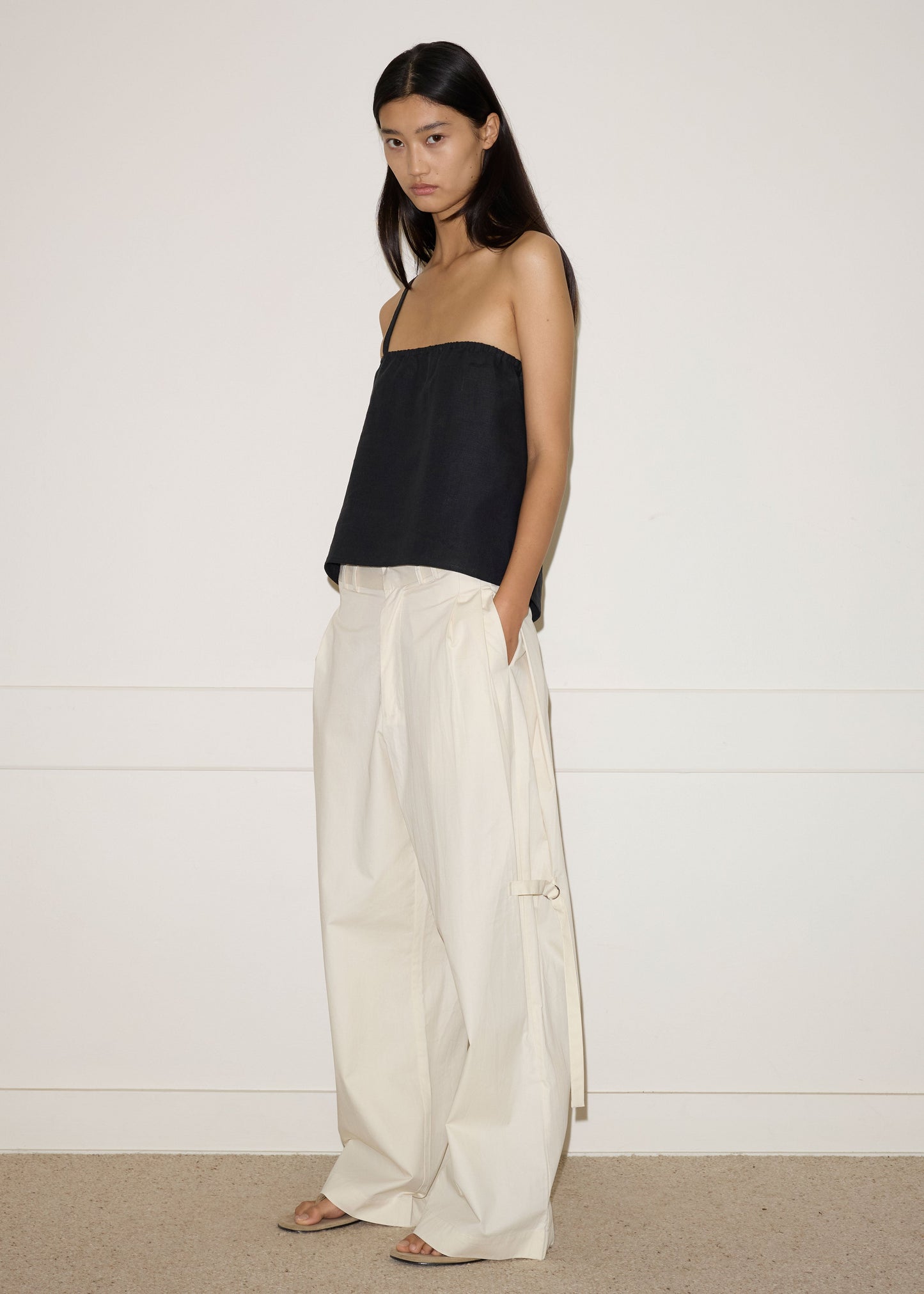A woman stands indoors against a plain white wall, wearing a black strapless top and Deiji Studios’ Tab Pant in off white—wide-leg, high-waisted organic cotton pants with side ties. She has long straight hair and a neutral expression.