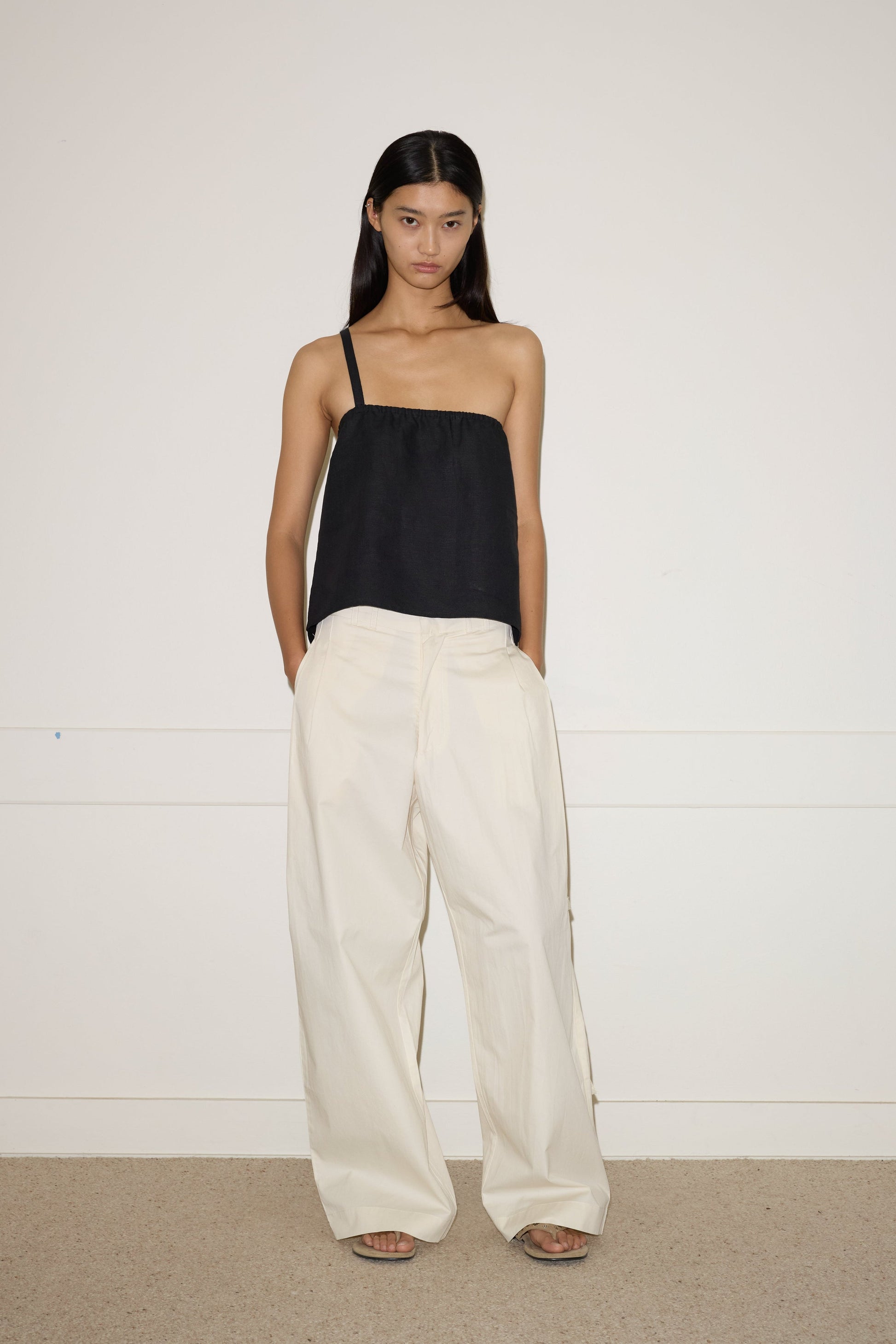 A woman with long, straight hair and a neutral expression stands against a white wall, wearing the 0 Tab Pant in Off White with her hands in her pockets, paired with a black top featuring an adjustable metal strap.