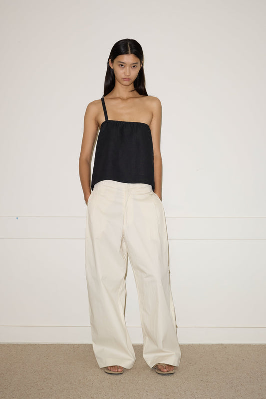 A woman with long, straight hair and a neutral expression stands against a white wall, wearing the 0 Tab Pant in Off White with her hands in her pockets, paired with a black top featuring an adjustable metal strap.