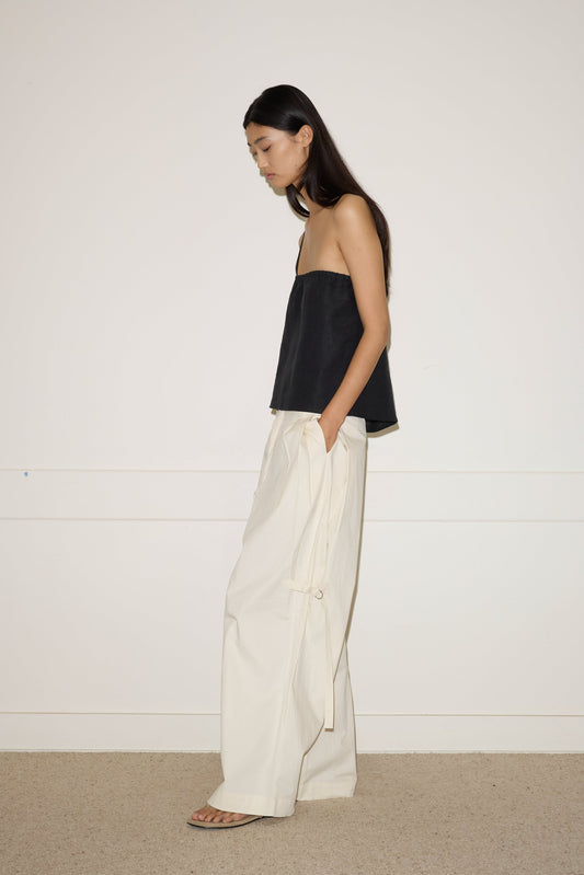 A woman with long dark hair stands in profile against a plain wall, wearing a strapless black top, the 0 Tab Pant in Off White with side ties, and flat sandals. Her hands are tucked into her pockets.