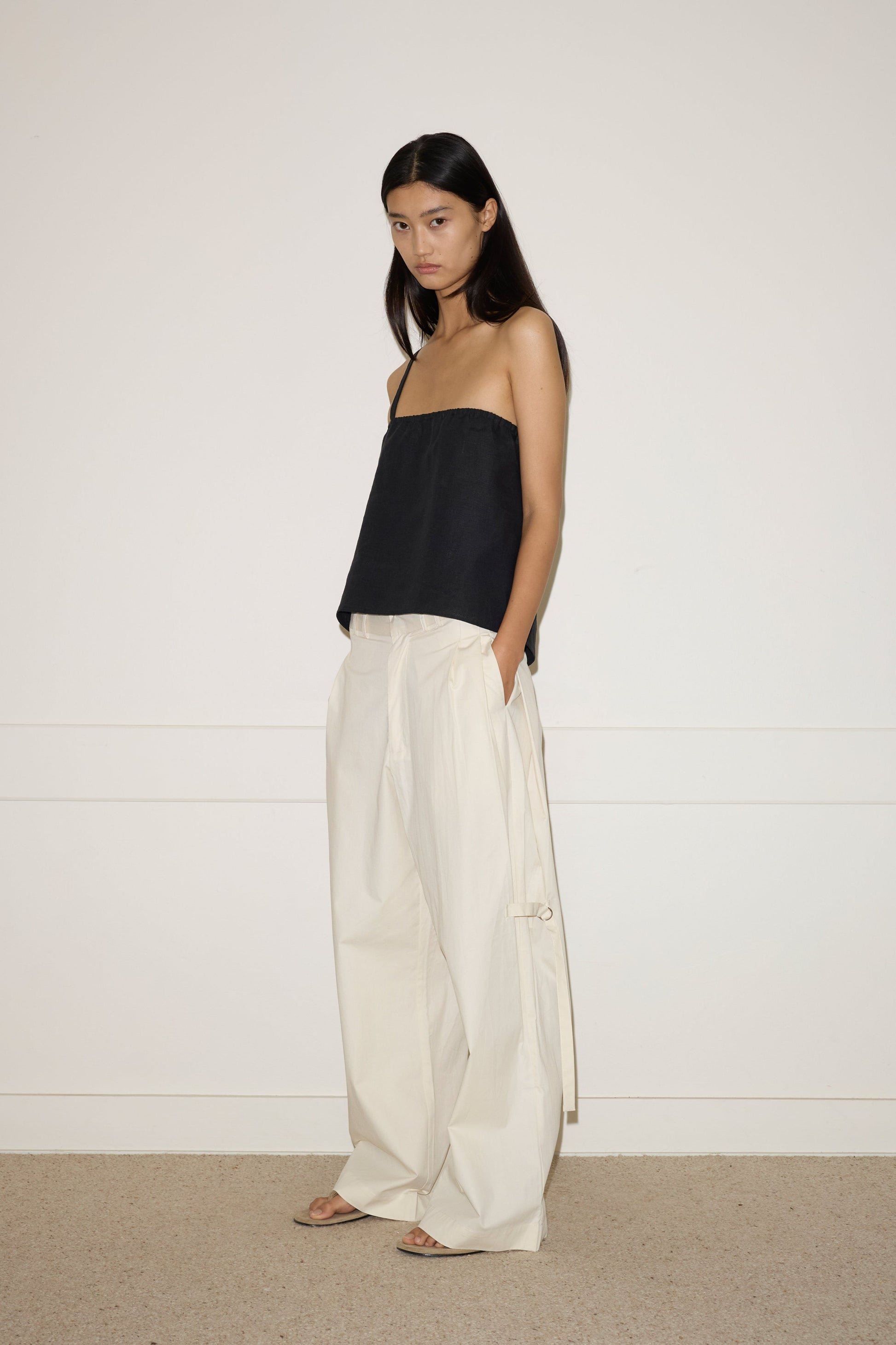 A woman with long, straight black hair stands against a white wall, wearing 0’s off-white Tab Pant—loose, high-waisted wide-leg trousers—with her hands in the pockets, paired with a black strapless top and open-toed sandals.